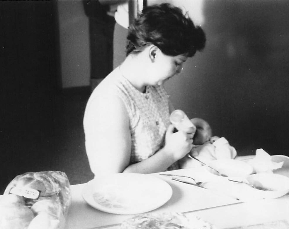 Woman feeding a baby with a bottle at a table with plates and bread nearby.