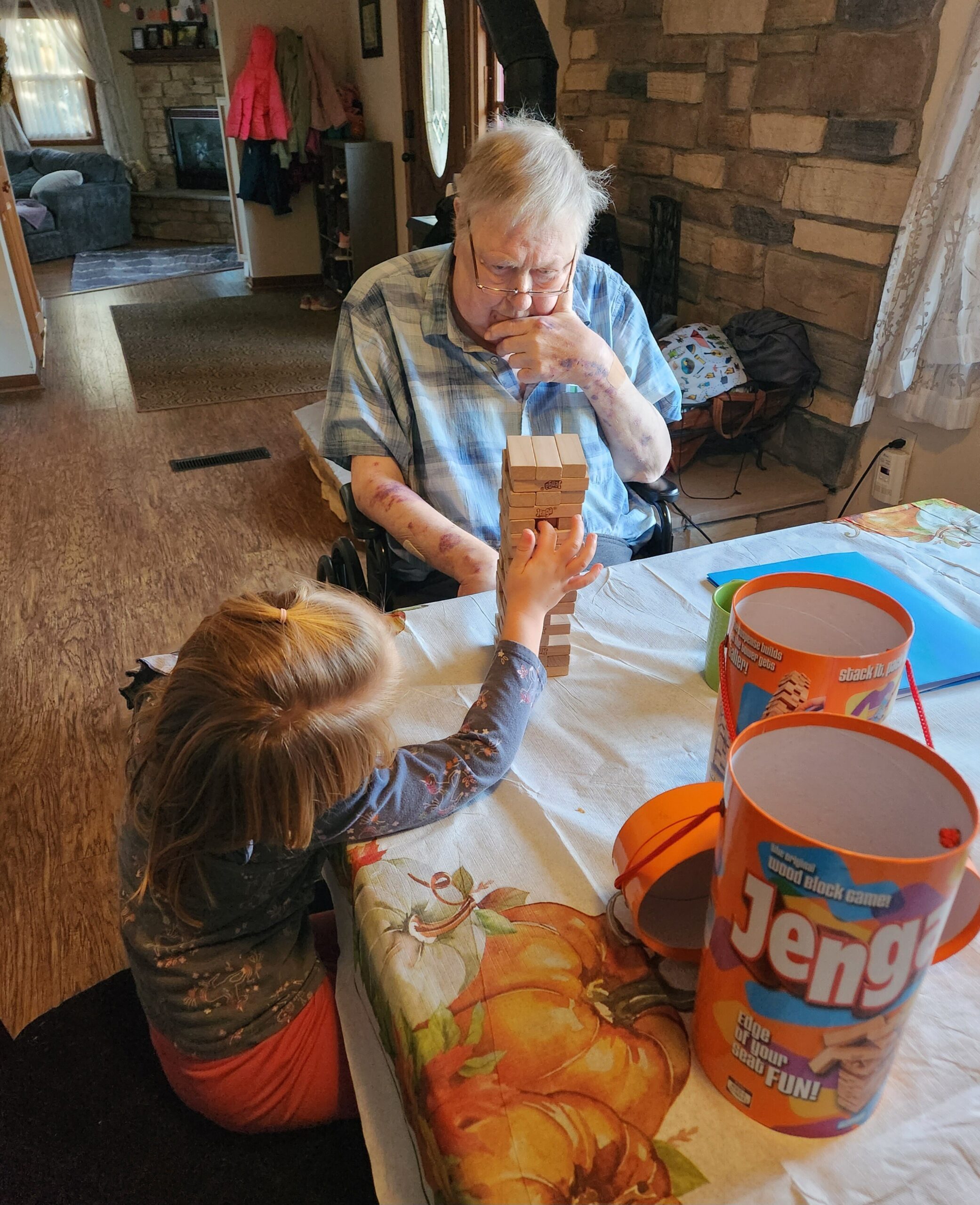 An elderly man in a blue shirt sits in a wheelchair, thoughtfully playing Jenga with a child at a dining table in a cozy living room with a stone wall.