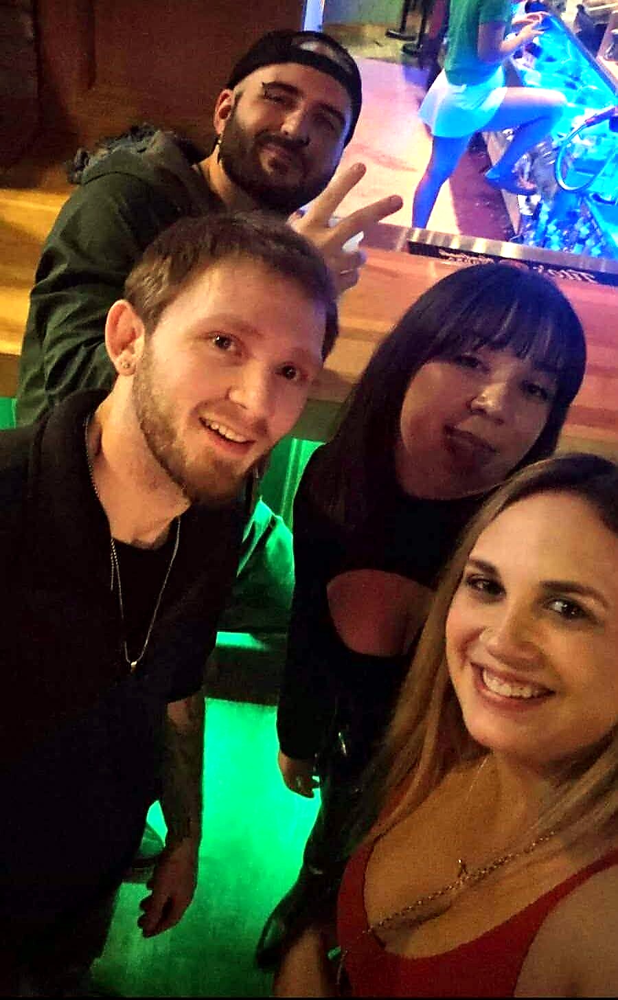 Four friends posing for a selfie in a nightclub with colorful lighting; two women in front and two men behind, one flashing a peace sign.