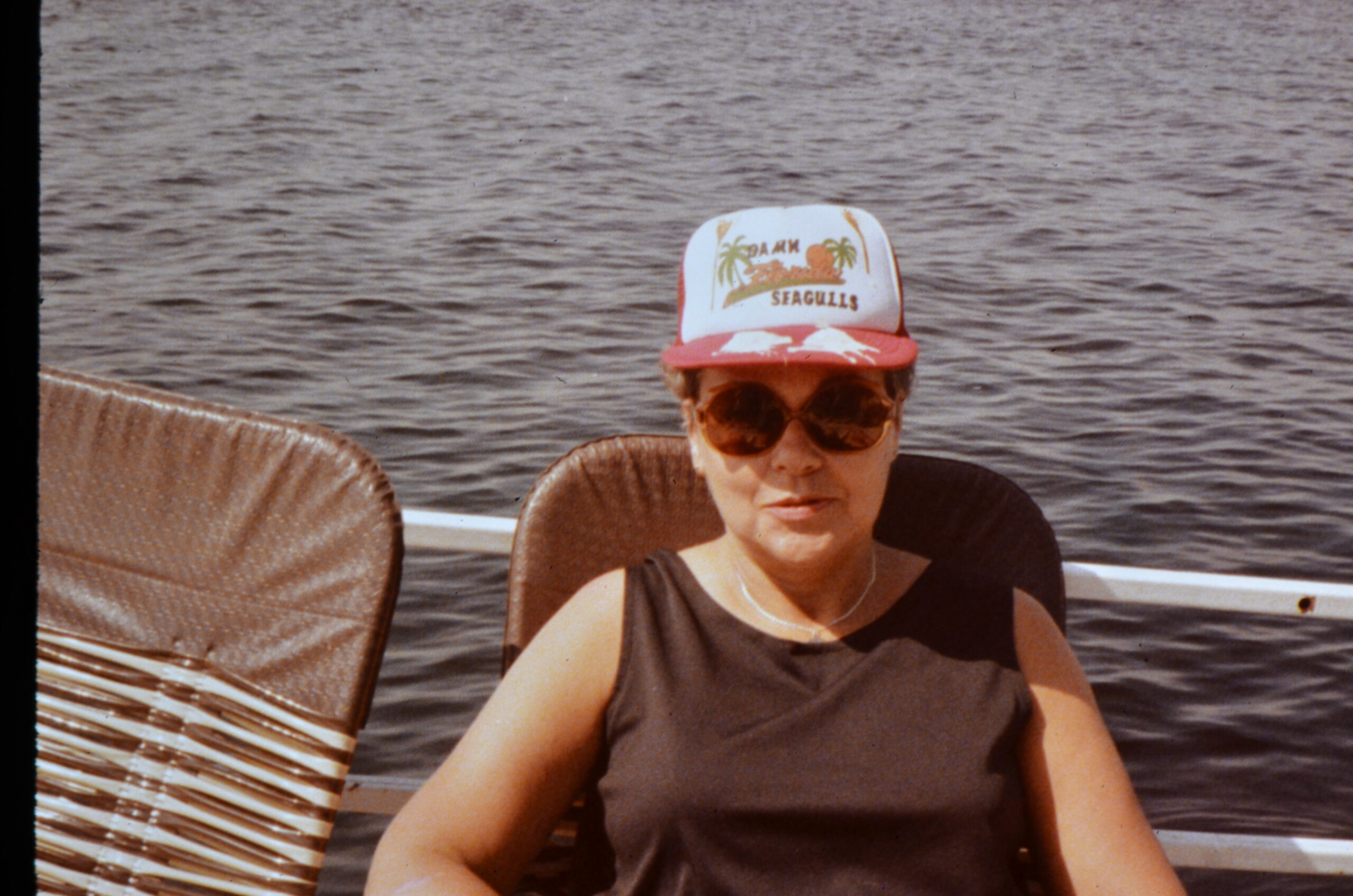 Person wearing sunglasses on a boat, white cap with palm trees and 'SEAGULLS' logo, brown water in the background.