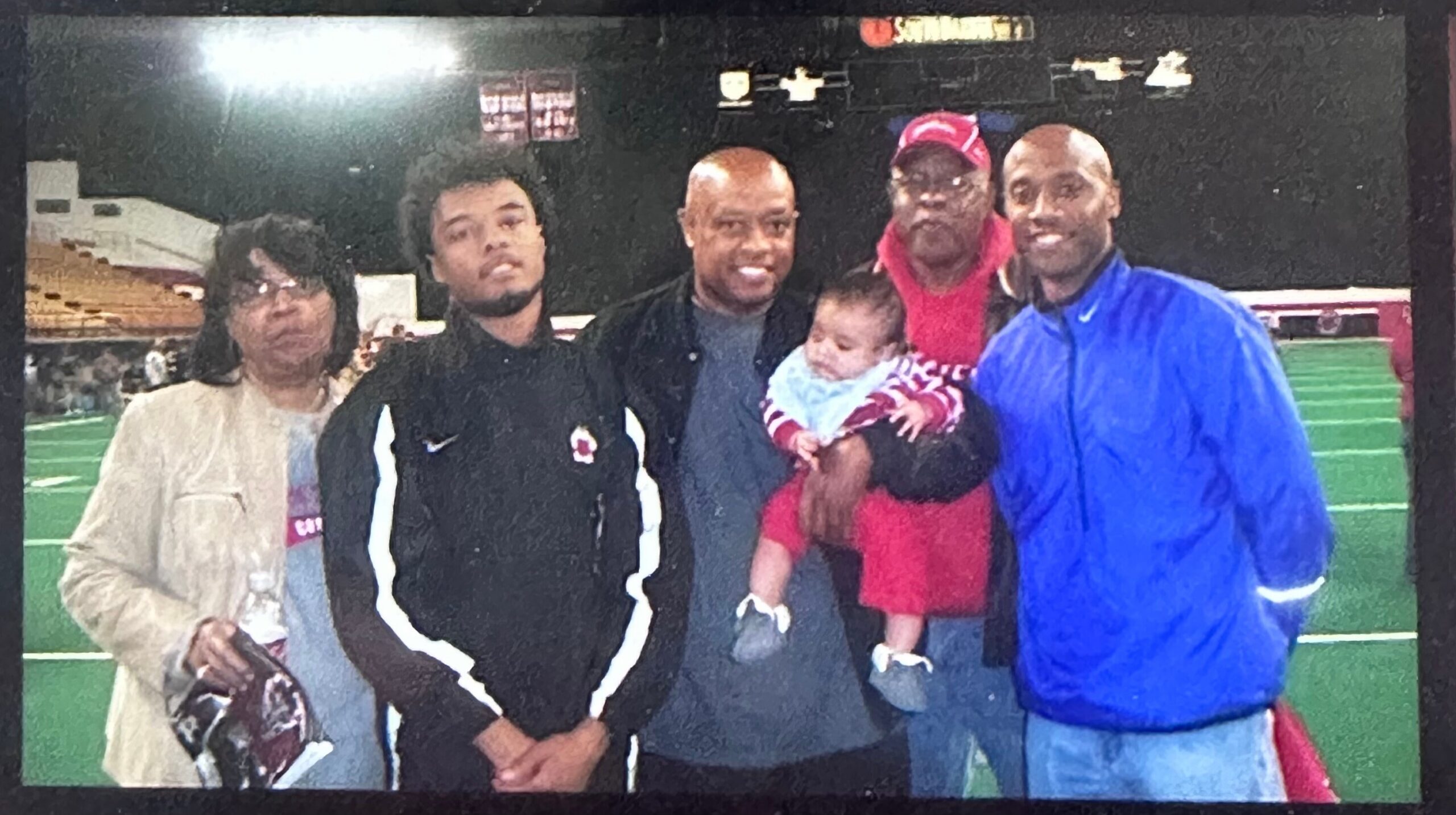 Group of six adults and a baby posing for a photo on an indoor sports field with artificial turf in the background, smiling at the camera as a team gathers.