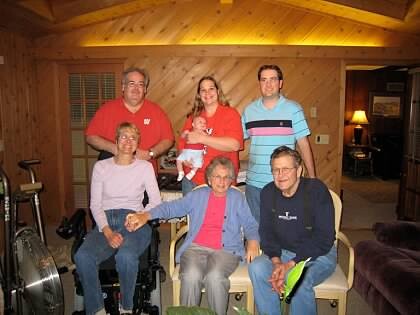 Seven people posing for a casual family photo in a wood-paneled living room with warm lighting, some seated front and others standing behind, including a baby.
