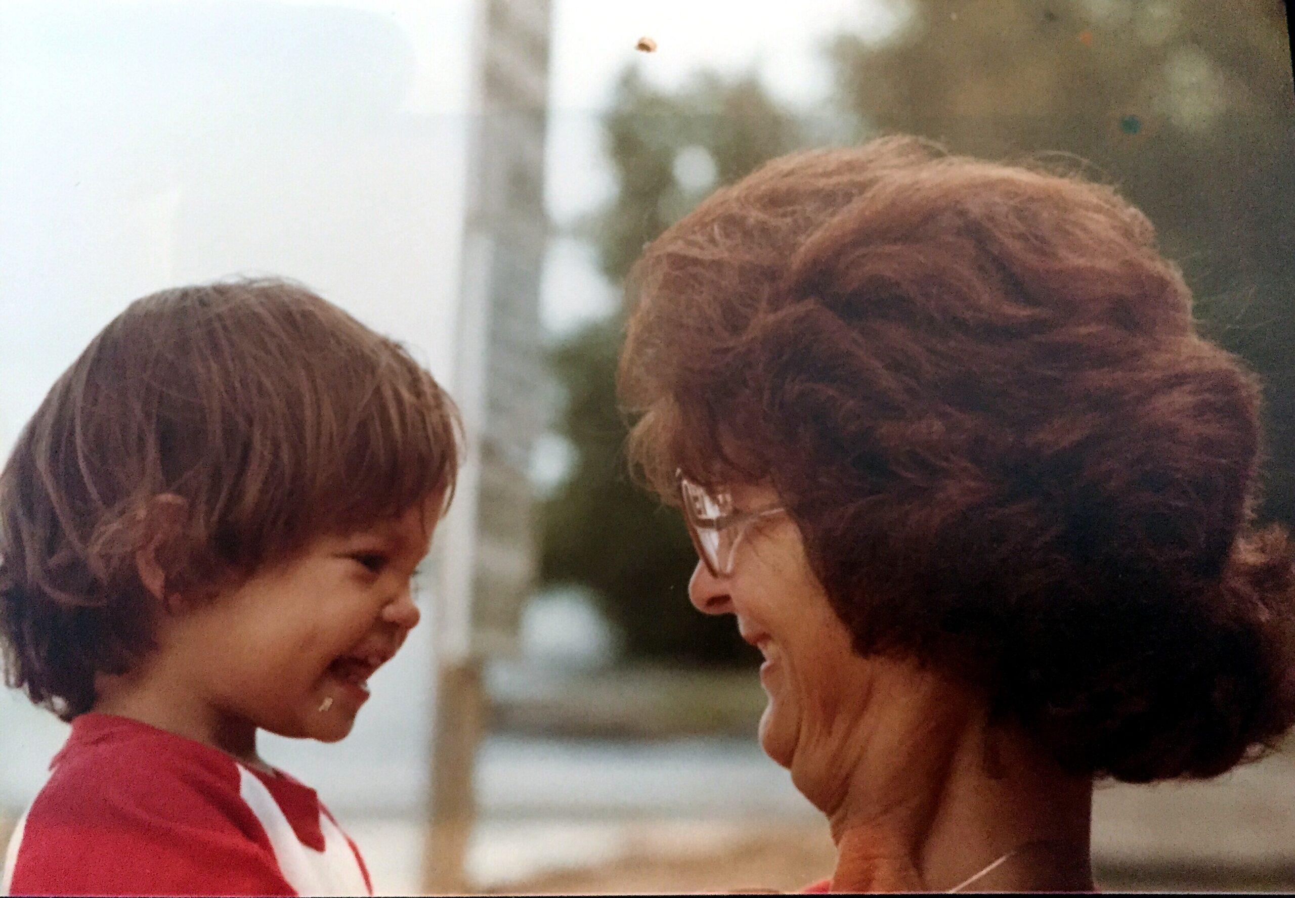 Two people smiling at each other in a close-up outdoor shot, a child in red on the left and an adult with brown hair on the right.