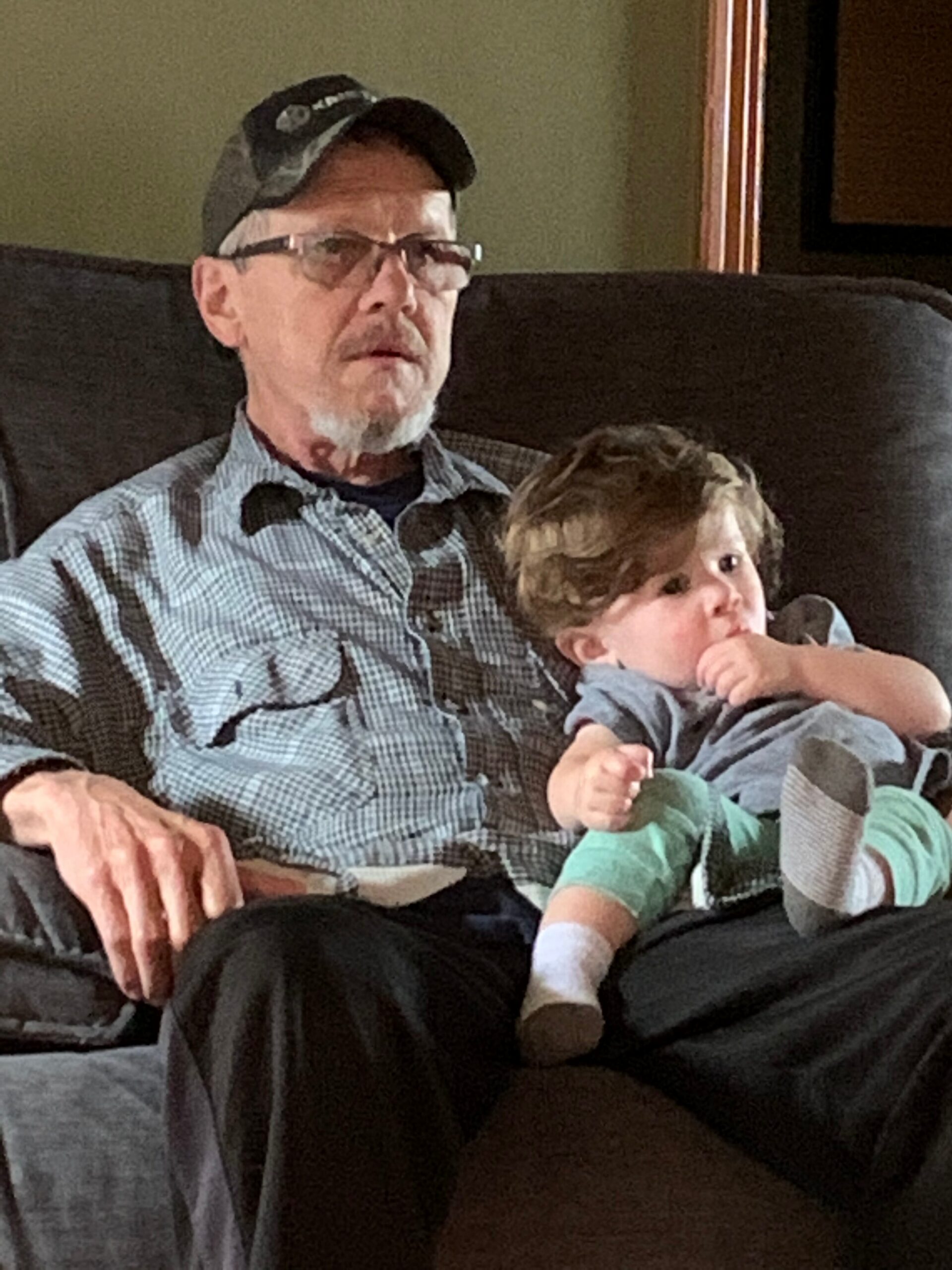 Older man with glasses and a cap sitting on a sofa, holding a young child on his lap in a living room setting.