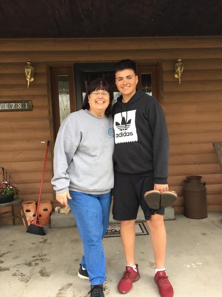 Two people smiling on a porch in front of a wooden house: a woman in a gray sweatshirt and jeans and a teen in a black Adidas hoodie, holding shoes.