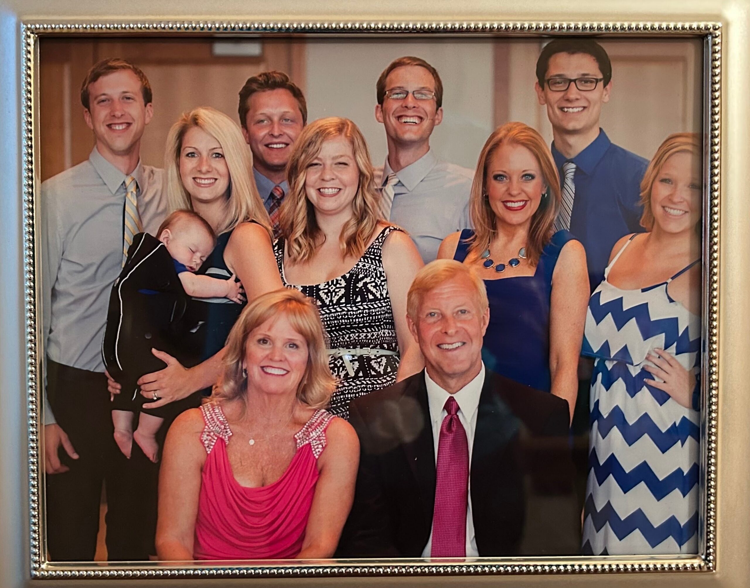Group portrait of friends and family in formal attire posing together, with a baby being held by one person, all smiling in a framed photo.