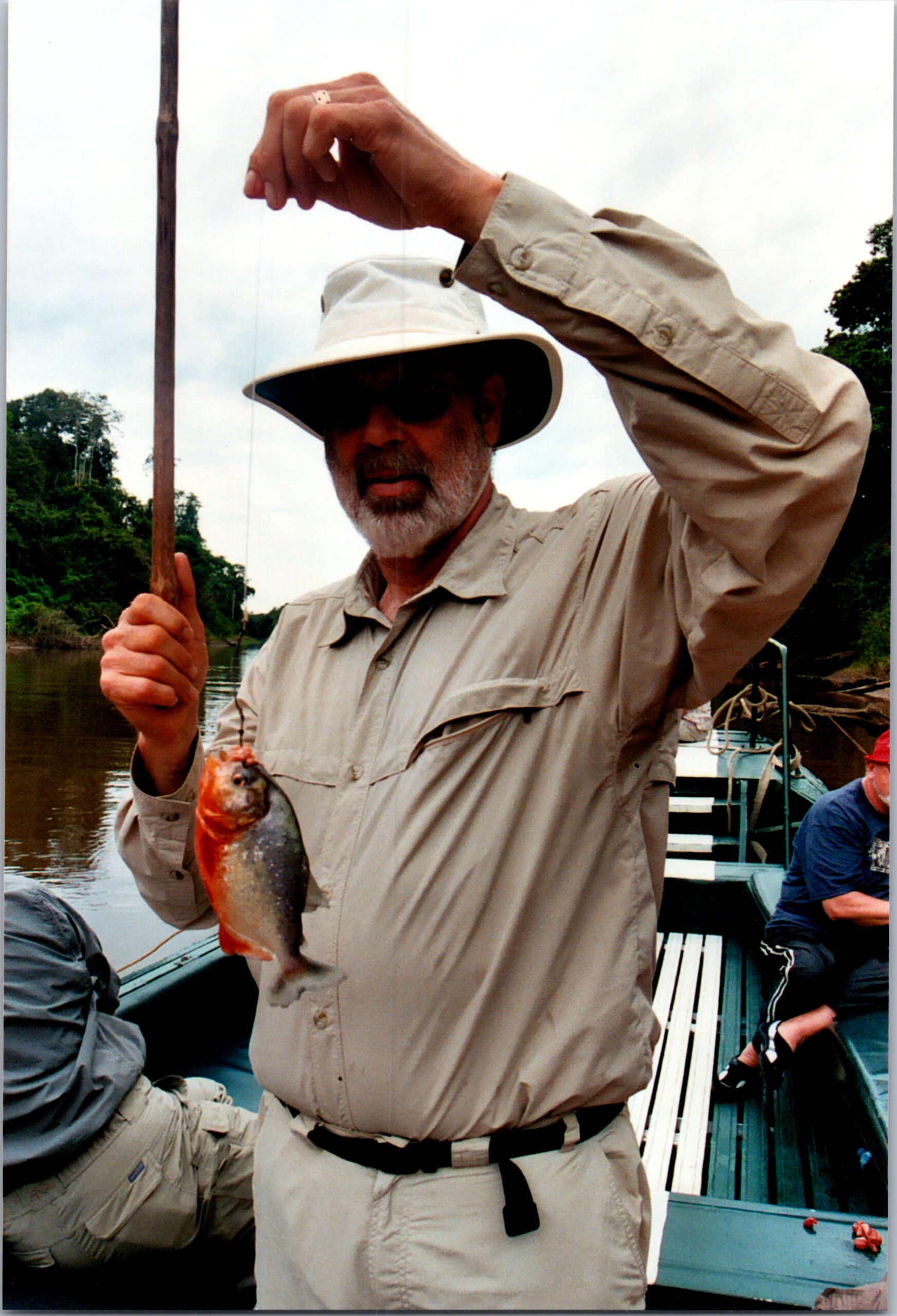 Man in a wide-brimmed hat and khaki shirt on a boat, holding up a colorful fish by the line to show his catch.