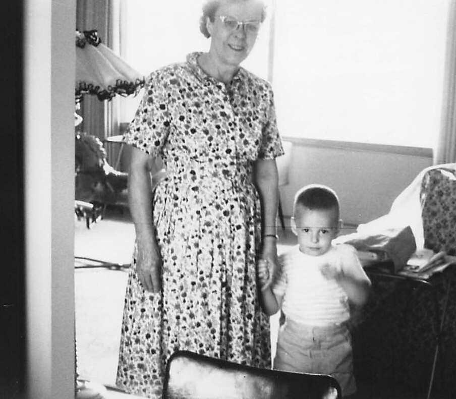 Older woman in a floral dress holding a young child's hand in a living room, both looking toward the camera.
