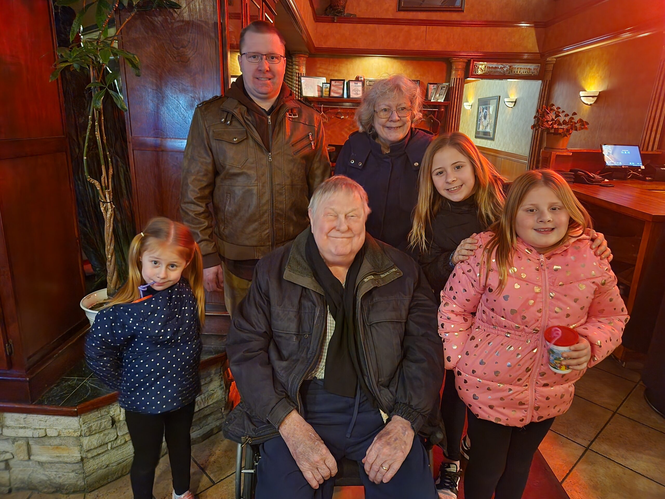 Group of five people posing for a photo inside a warmly lit restaurant, including an elderly man in a wheelchair and smiling family members nearby.