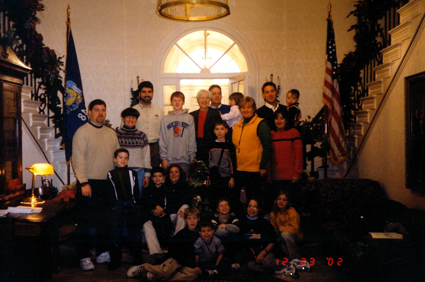 Group of adults and children posing for a photo in a formal, flag-flanked lobby with a curved staircase in the background