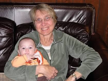 Smiling older woman with short gray hair sitting on a dark leather sofa, holding a baby in her lap.