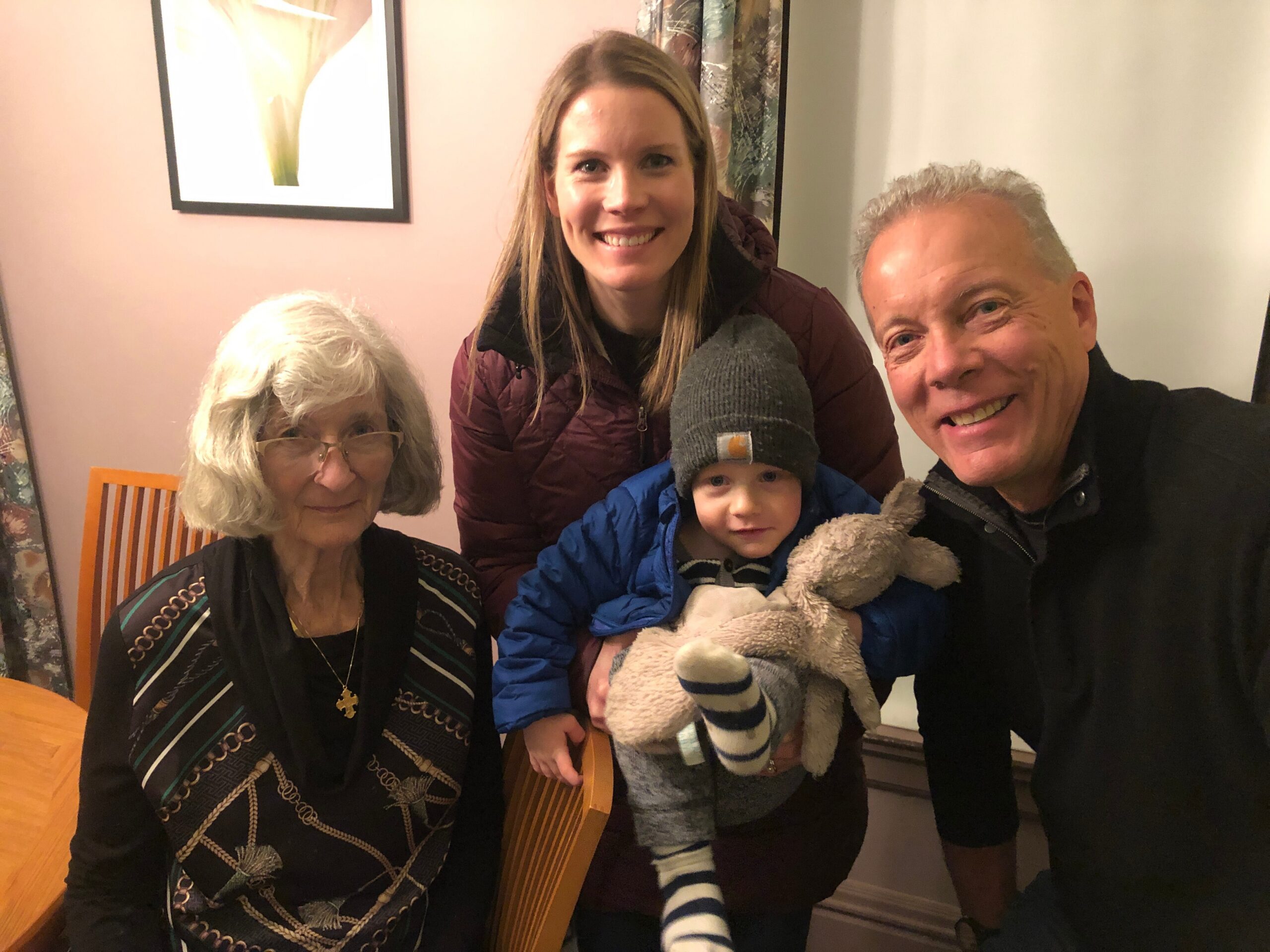 Smiling family taking a selfie indoors: elderly woman on the left, smiling young woman center, child in blue jacket with a striped hat holding a teddy bear, and a man on the right.