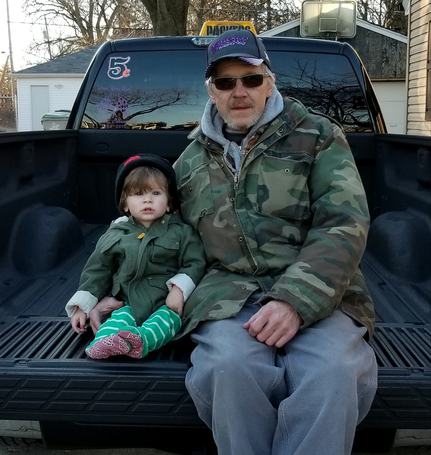 An older man in a camo jacket and cap sits with a young child in the bed of a pickup truck, outdoors.