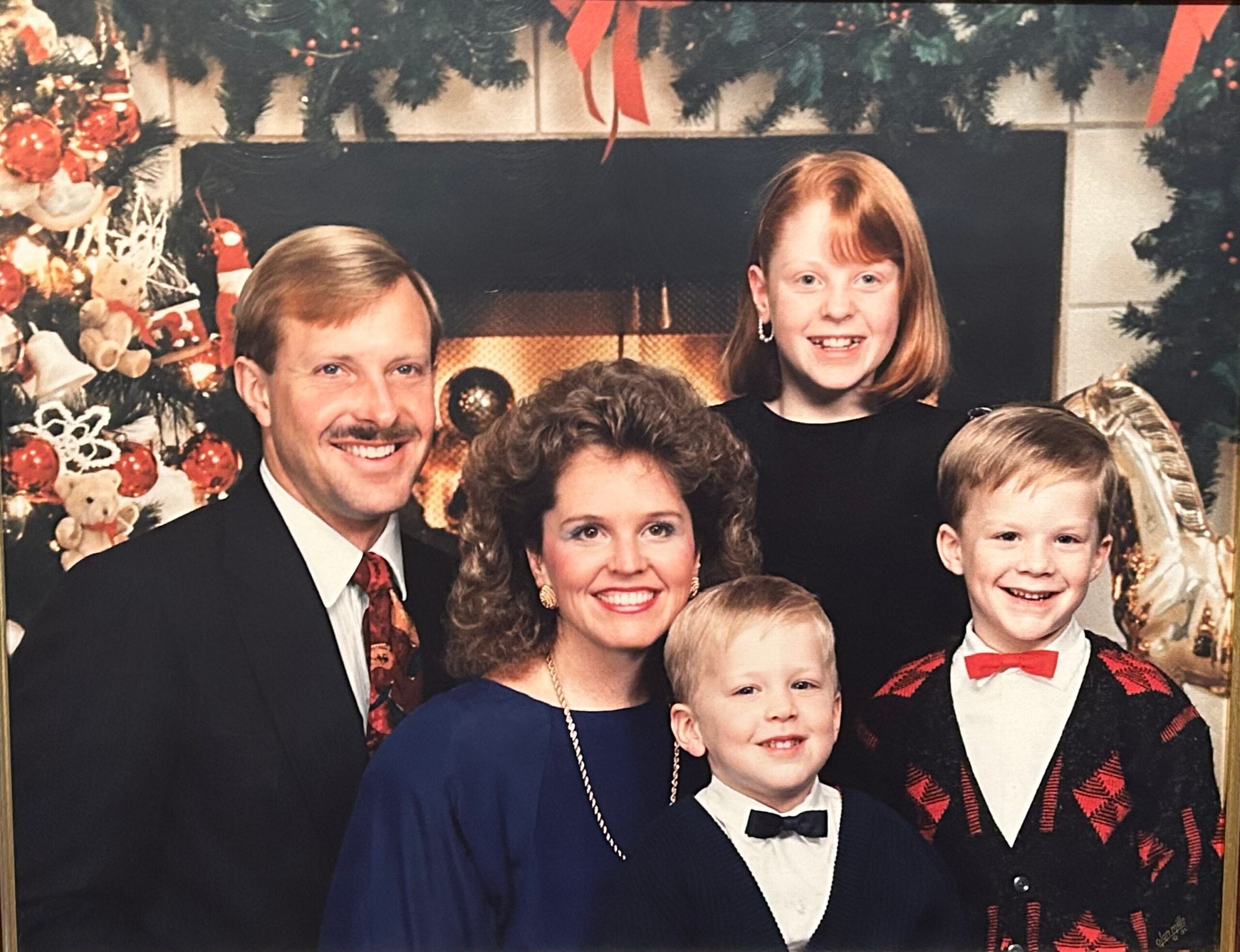 Family portrait by a decorated Christmas tree and fireplace; adults and children in formal attire smiling.