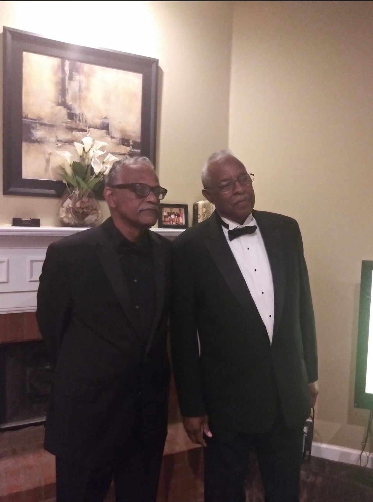 Two elderly men in tuxedos posing indoors, one with glasses, holding hands in front of a fireplace and a painting.