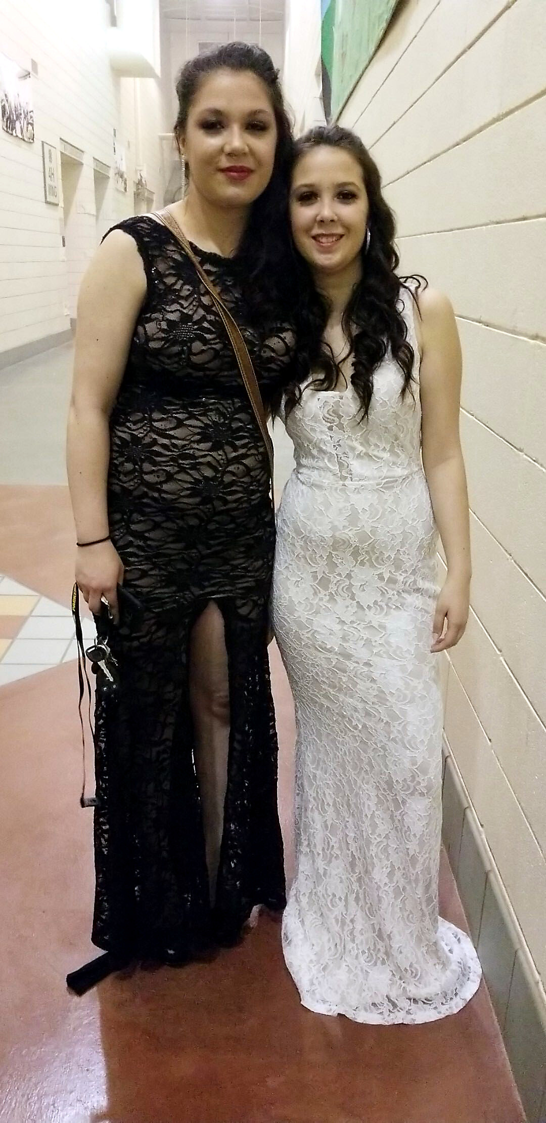 Two women in a hallway posing for a photo, one in a black lace gown with a slit and the other in a white lace dress, smiling together.
