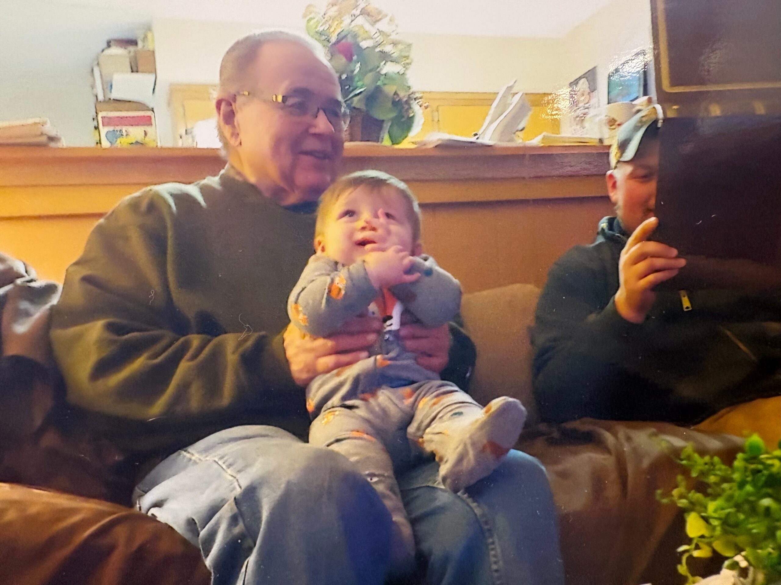 Older man holding a baby on a brown sofa in a living room, with another person partially visible on the right taking a photo or video with a device