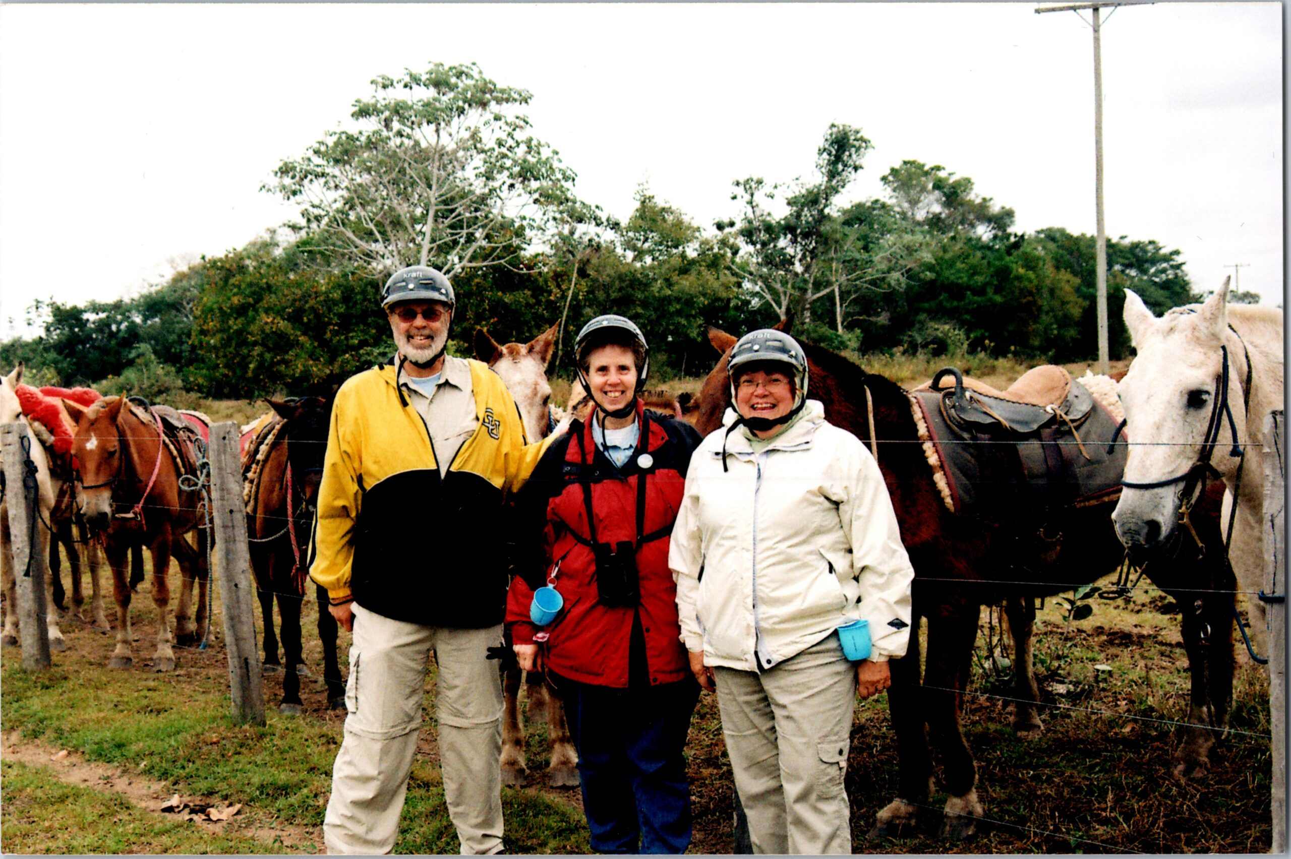 Three people in riding gear and helmets stand in front of saddled horses behind a wooden fence in a pasture.