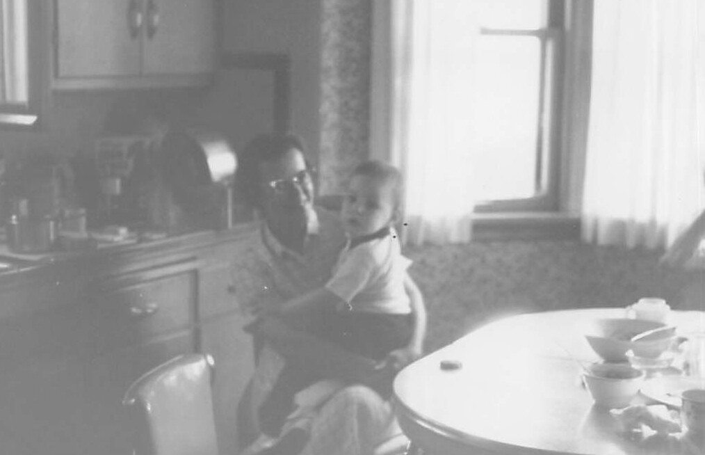 Man with glasses sits in a kitchen, holding a baby on his lap near a round table with dishes.