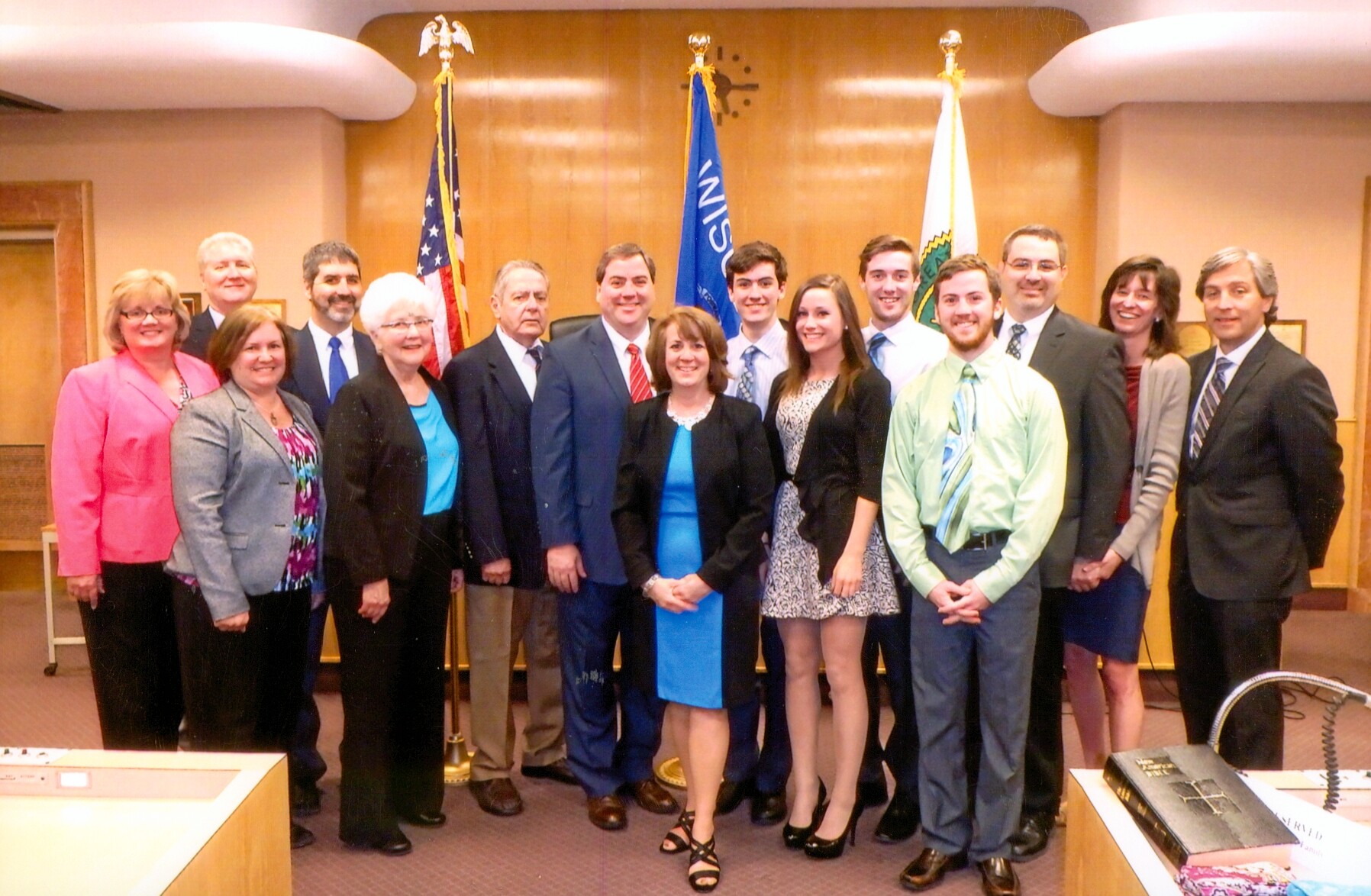 Group of professionally dressed people posing for a formal photo in a government meeting room with flags behind them.
