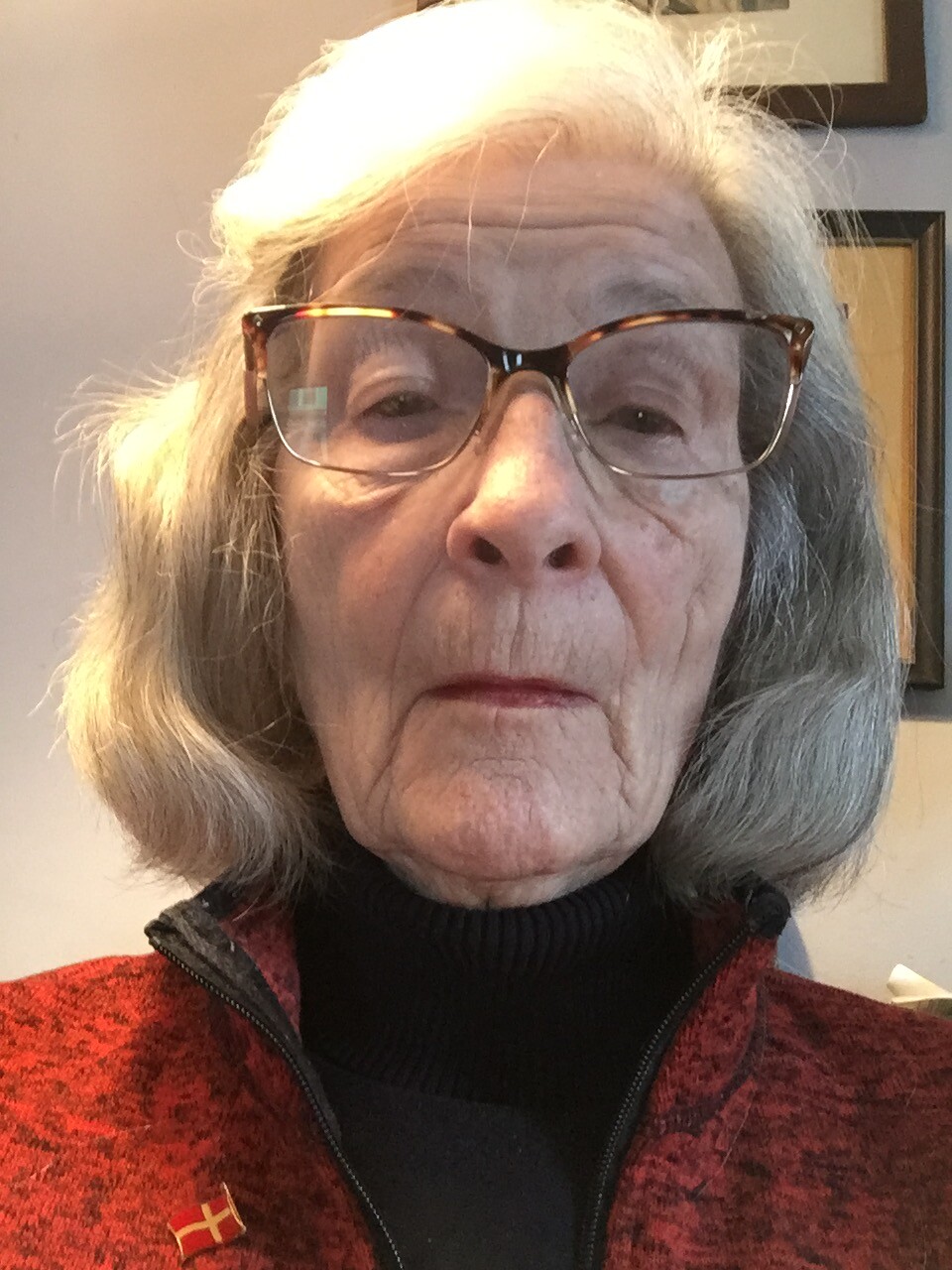 Portrait of an elderly woman with short gray hair and tortoiseshell glasses, wearing a red zippered sweater over a black turtleneck, with a Danish flag pin on her lapel.