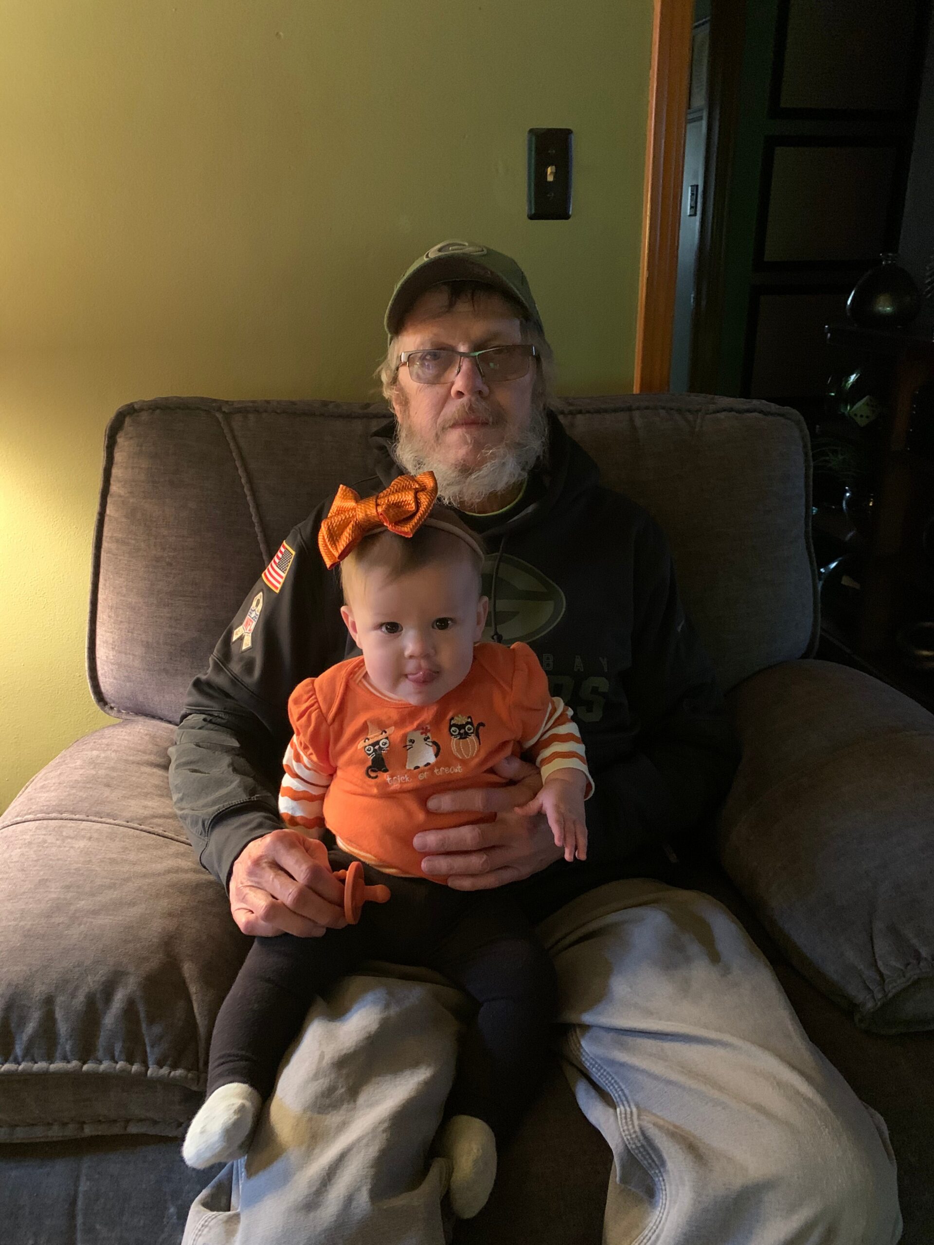Older man with a beard and glasses sits on a couch wearing a cap, holding a baby in an orange onesie with an orange bow headband on his lap.
