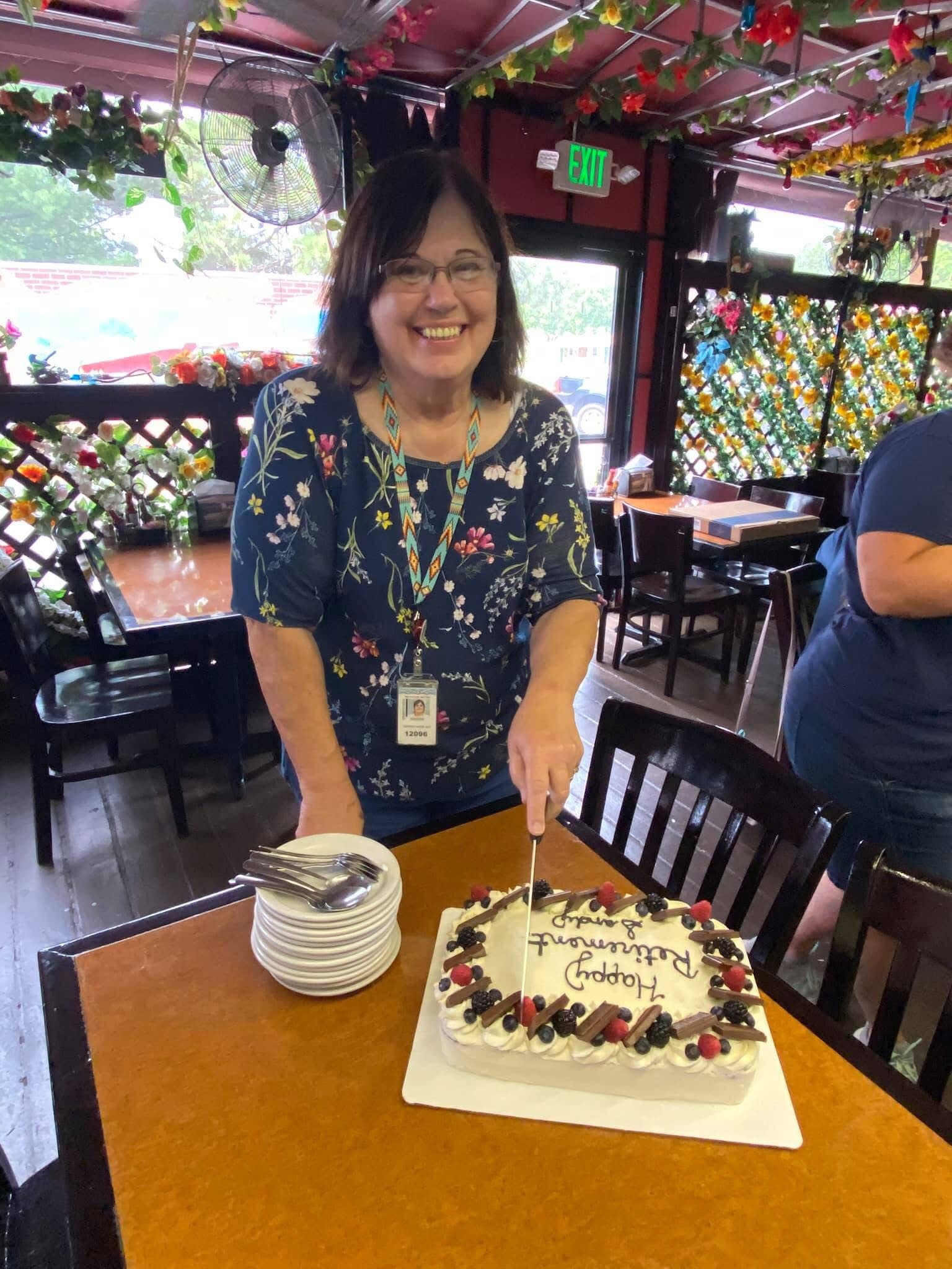 Smiling woman in a blue floral top cutting a rectangular birthday cake topped with berries at a decorated restaurant table.