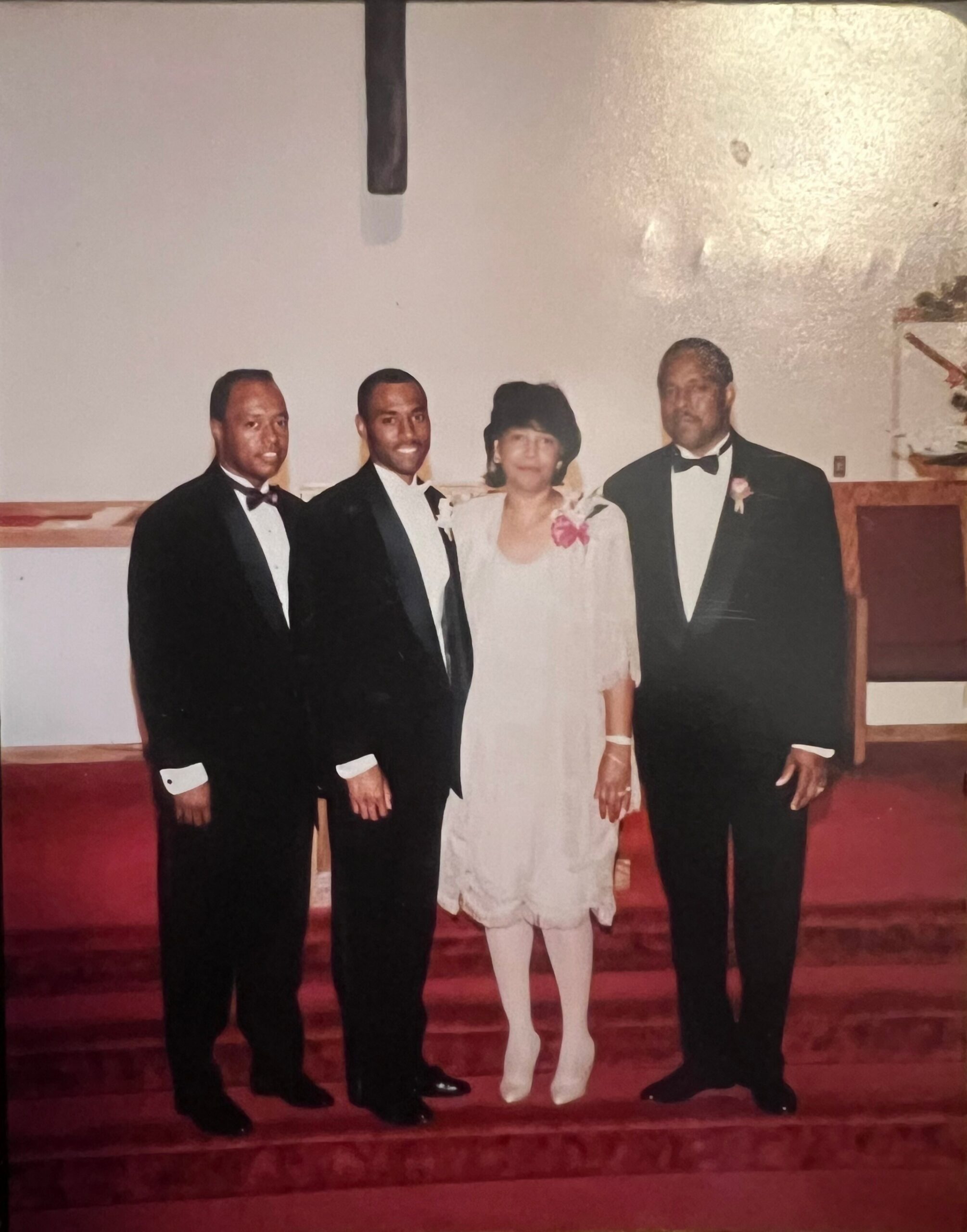 Four adults in formal attire pose together on red-carpeted church steps, with a woman in a light dress flanked by three men in tuxedos, likely at a formal event or wedding.