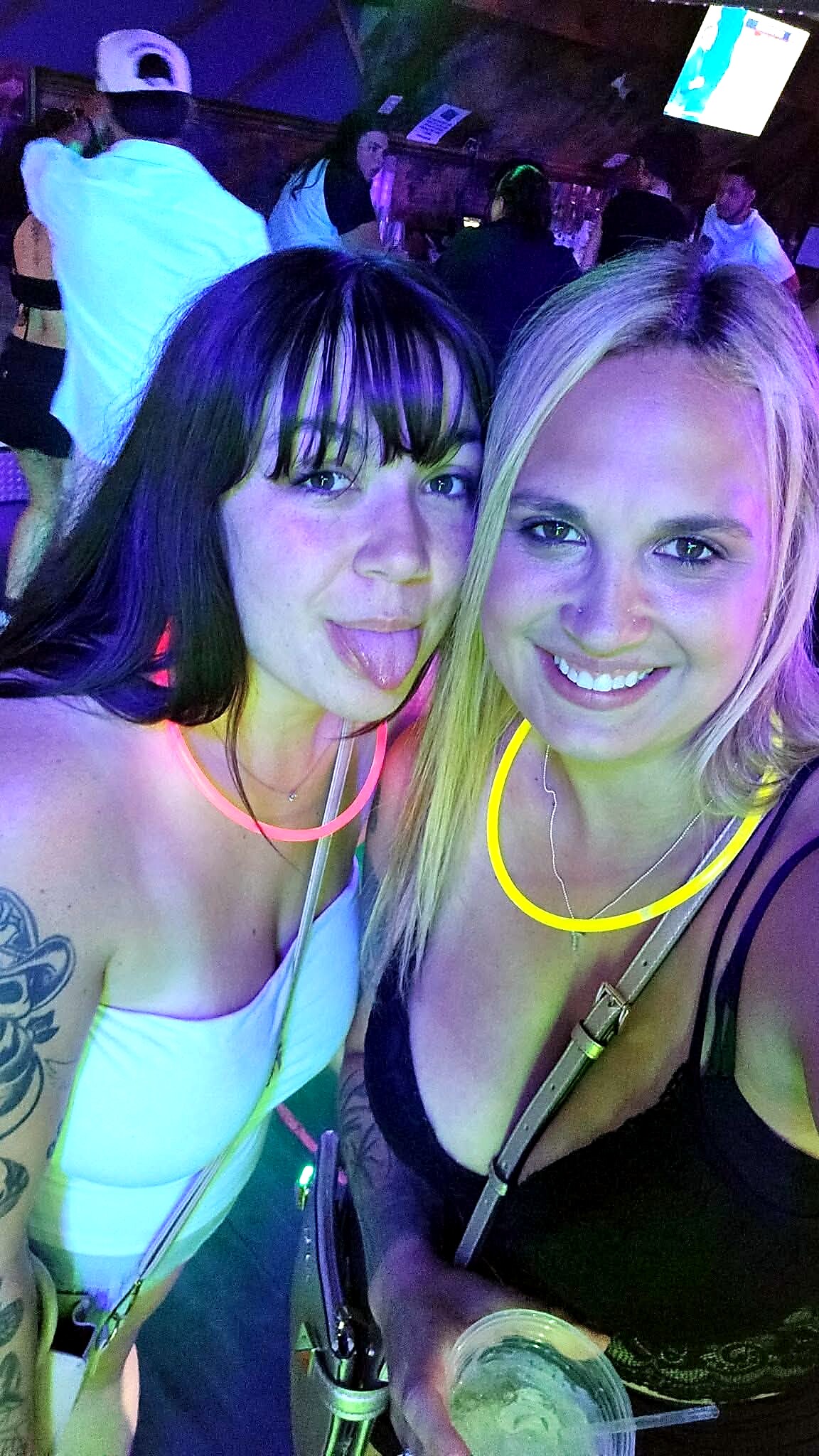 Two women at a nightclub taking a selfie, wearing glowing neon necklaces and smiling with drinks in hand.