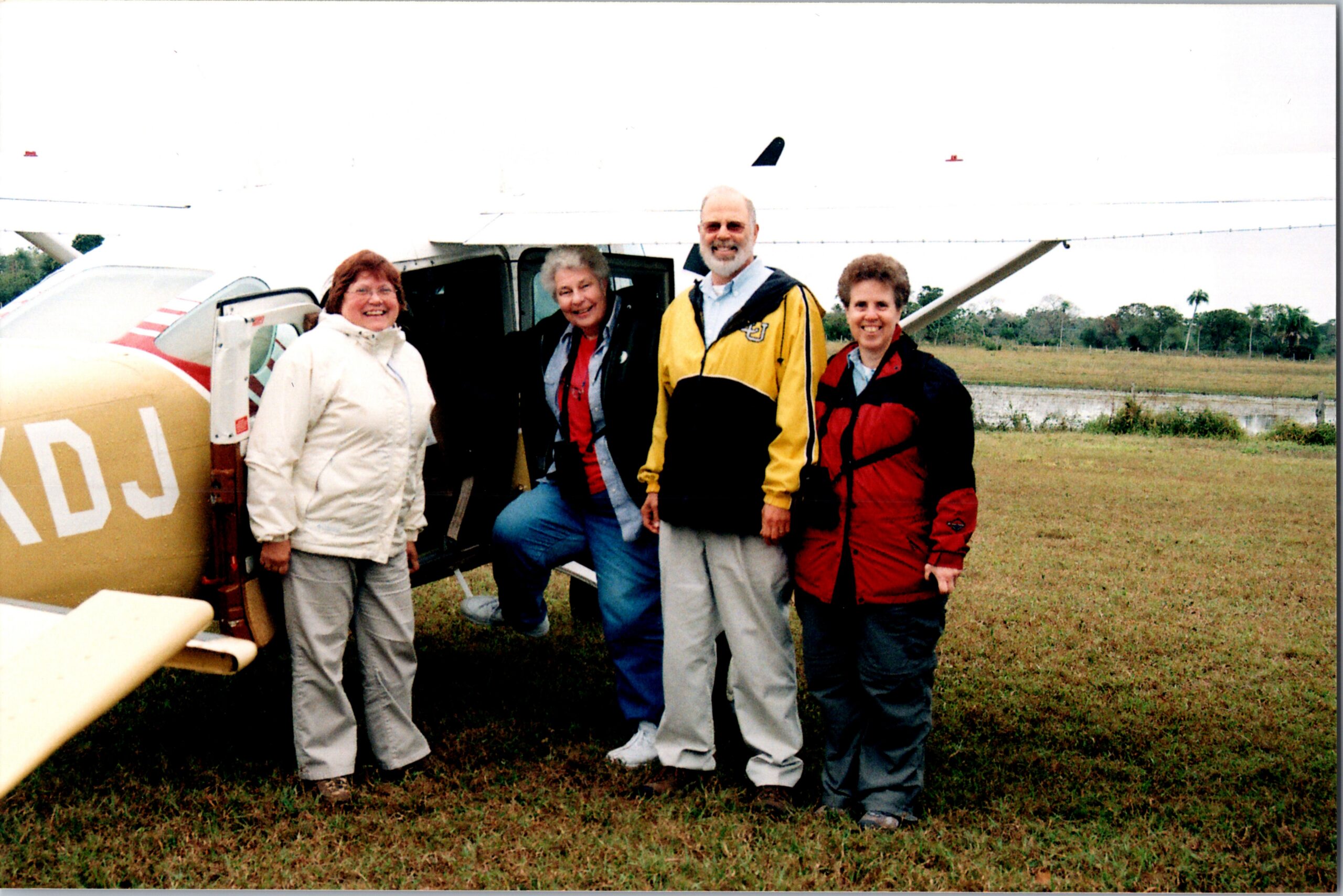 Four adults stand beside a small yellow airplane on a grassy airfield, smiling at the camera with the plane door open behind them.