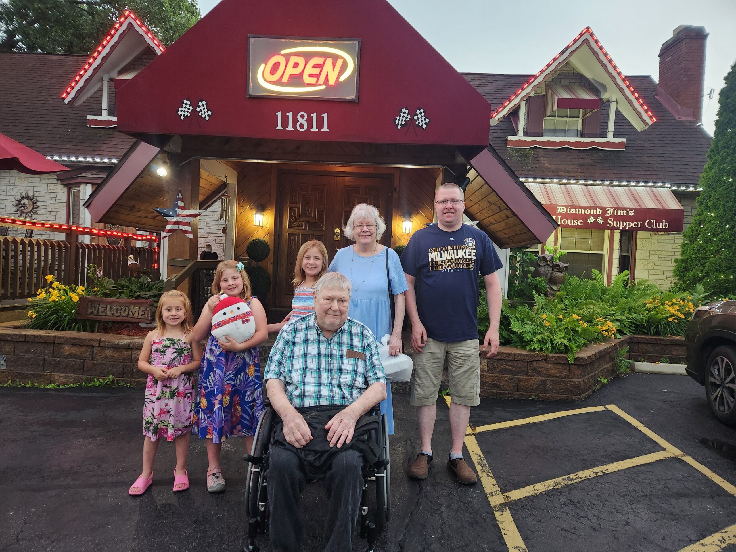 Family group photo outside a restaurant entrance with a red roof and open sign in background; smiling adults and children posing together, one person in a wheelchair foreground.