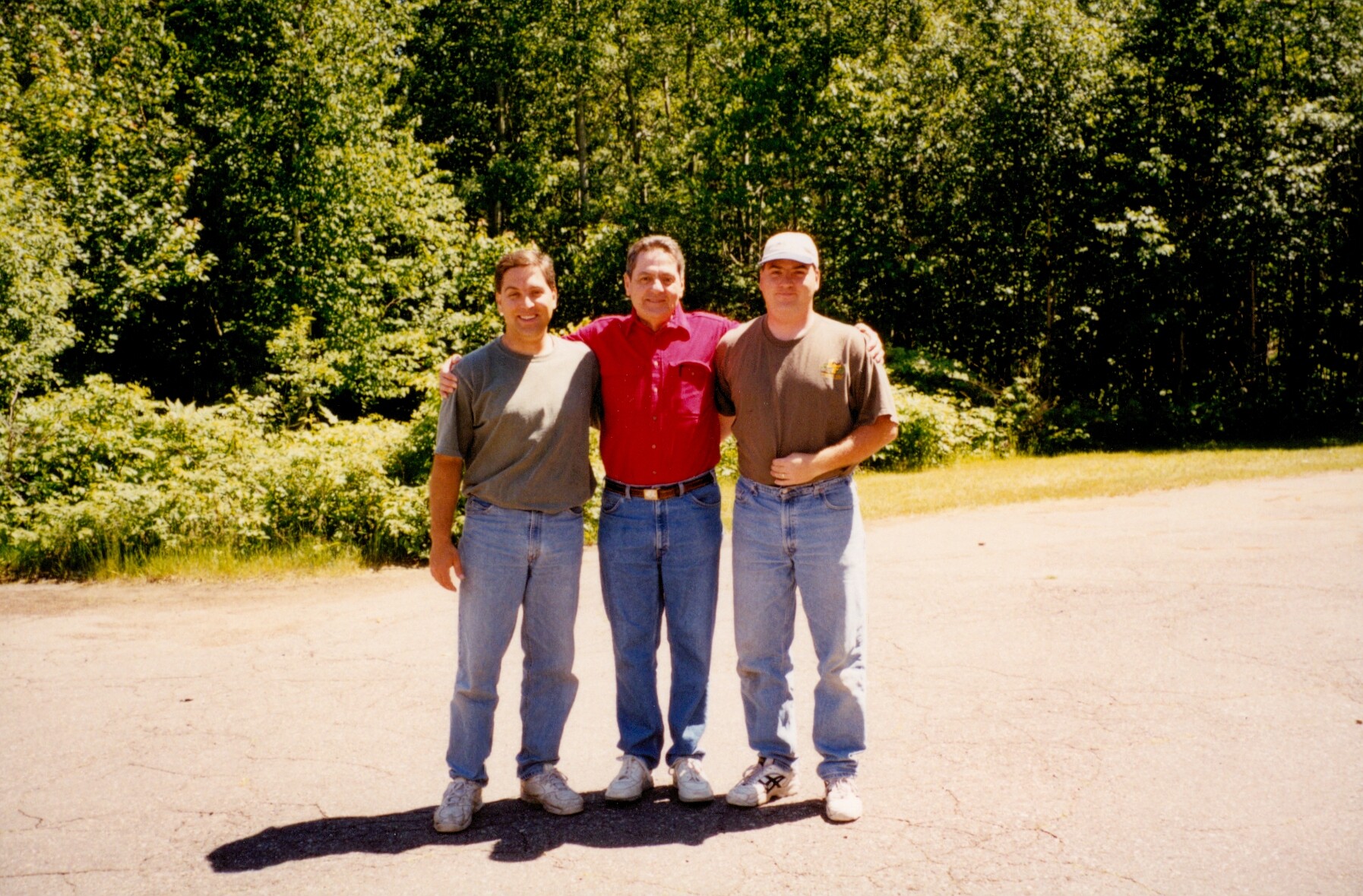 Three men in casual clothes pose together outdoors, arms around each other, on a sunny day with trees in the background.