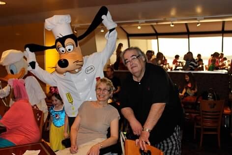 Goofy-like cartoon dog mascot in a chef outfit poses with an elderly couple at a busy restaurant.