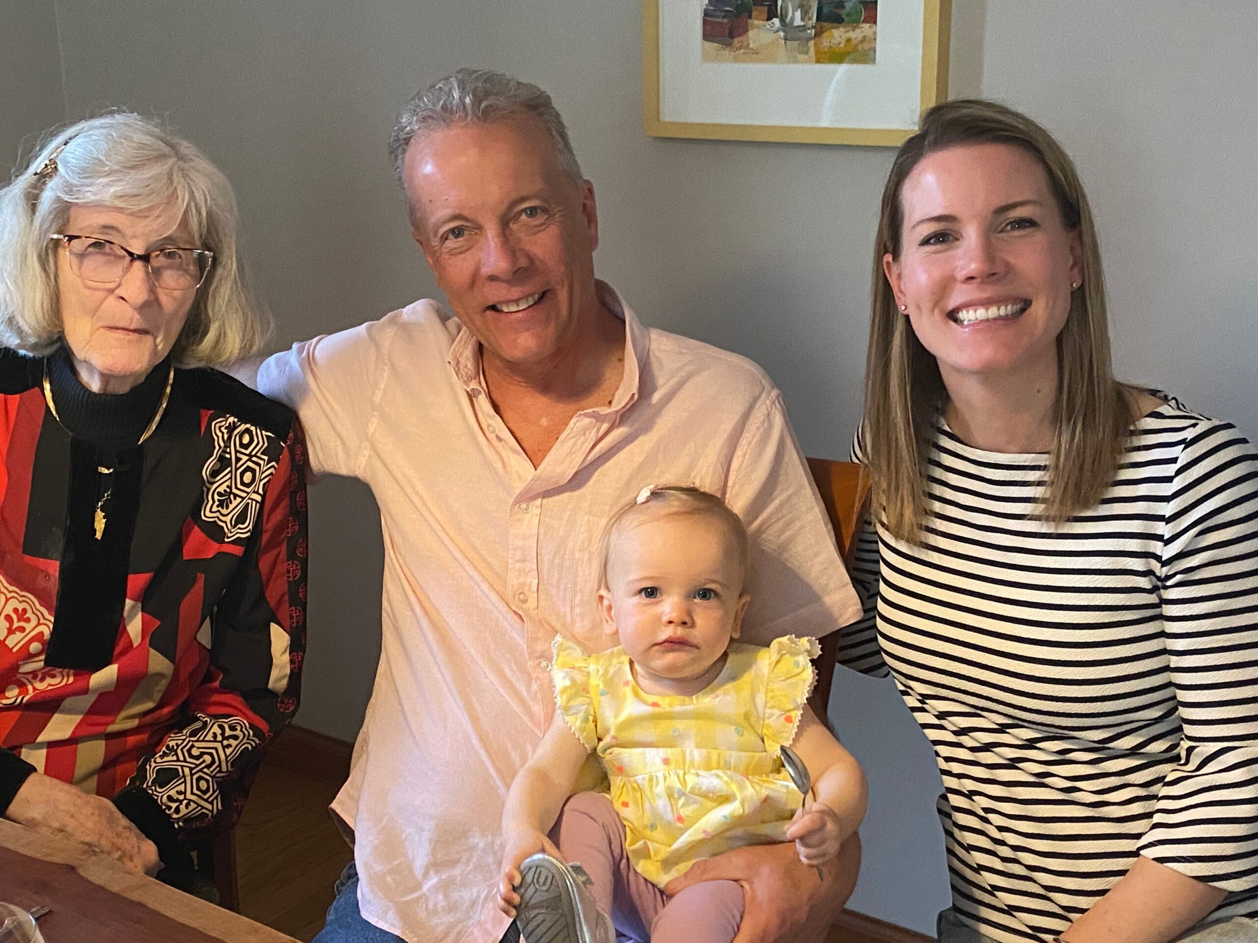Smiling grandmother, man, young woman, and baby girl seated together at a table, posing for a family photo.