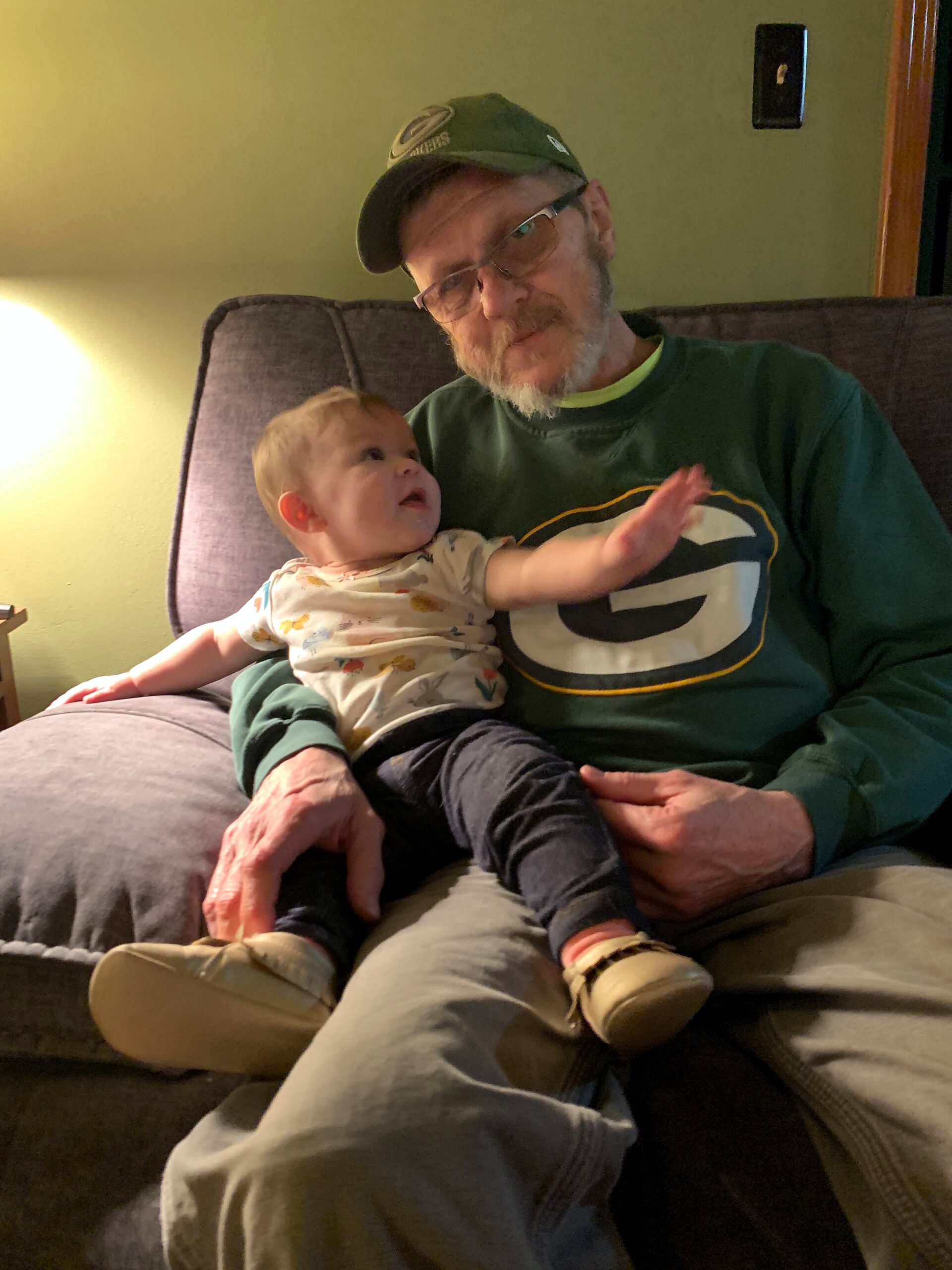 Older man with glasses and a green cap holding a small child on a sofa, looking at the baby.
