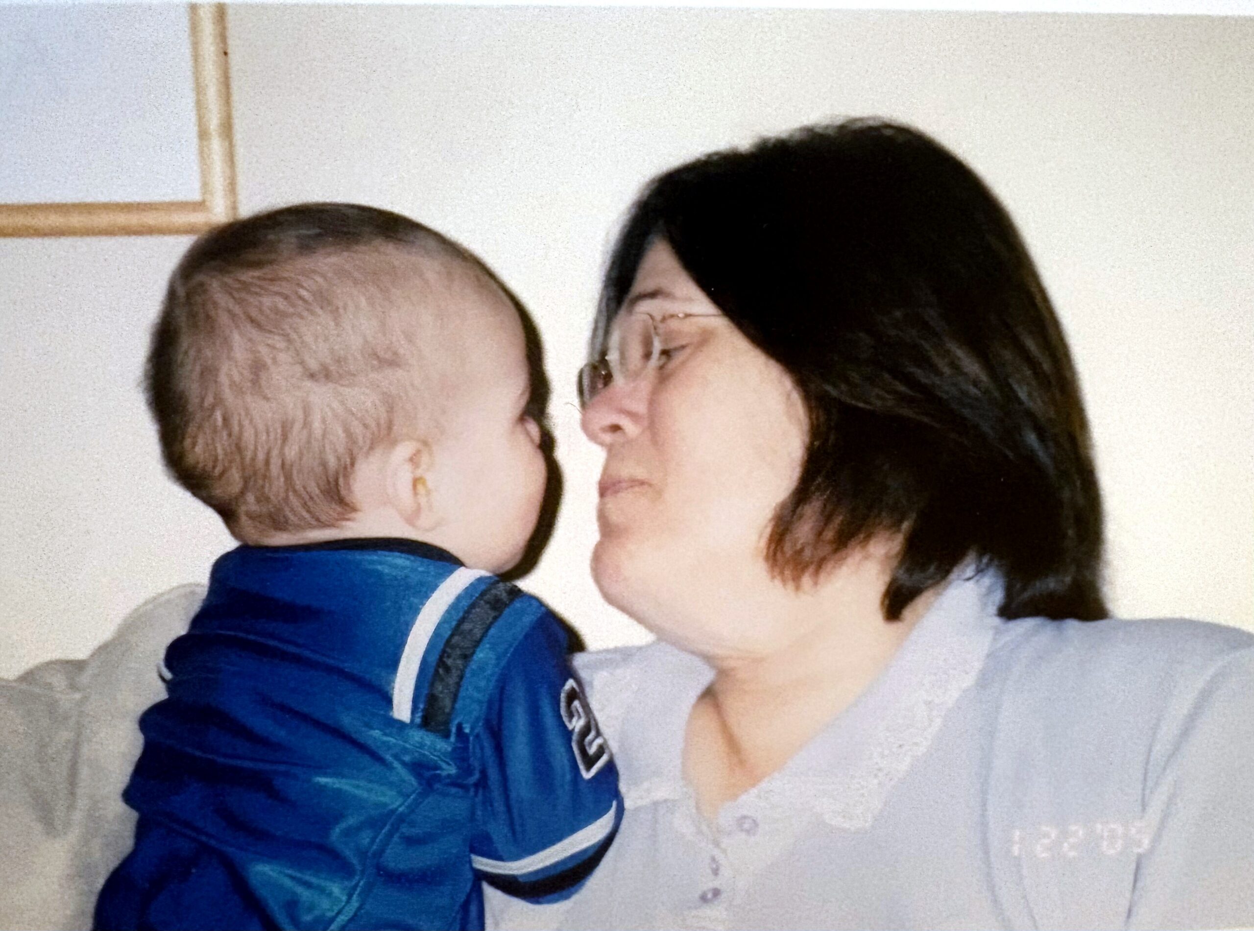 Woman with short dark hair and glasses leaning in to touch noses with a smiling baby wearing a blue striped shirt indoors.