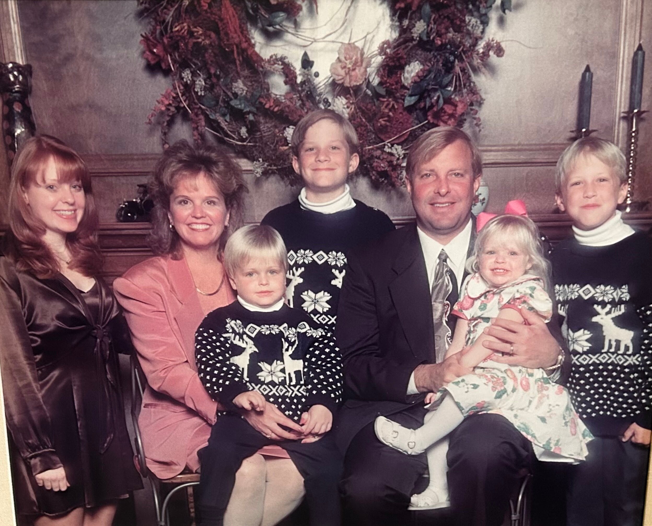 Family group portrait: parents with four children in festive sweaters, seated on a sofa in front of a Christmas wreath.