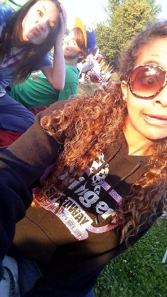 Young woman in the foreground wears large sunglasses and smiles during a sunny outdoor group selfie; others in colorful shirts in the background.