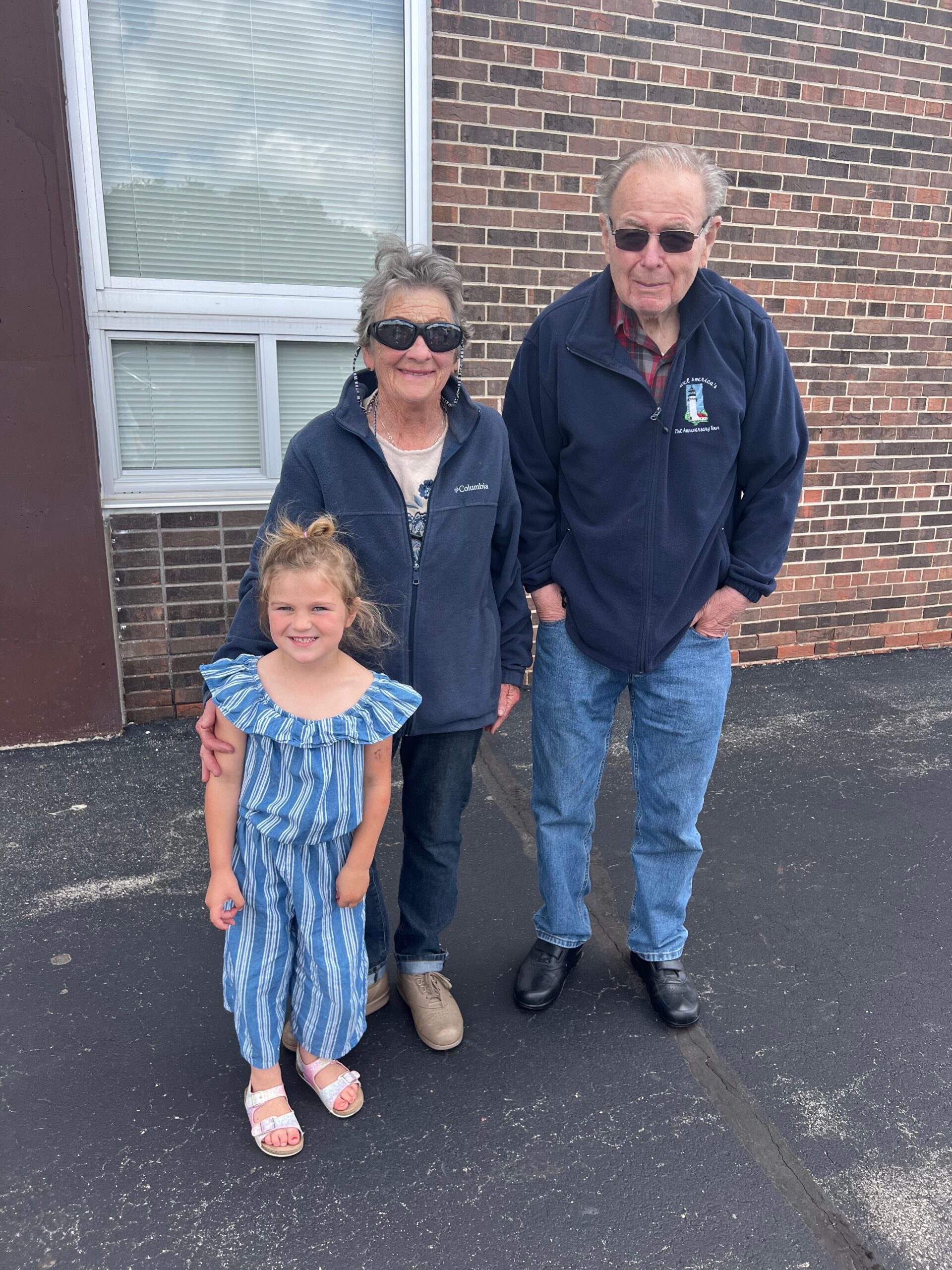 Three generations posing outside a brick building: an elderly couple with a young girl in a blue striped dress, all wearing casual jackets and sunglasses.