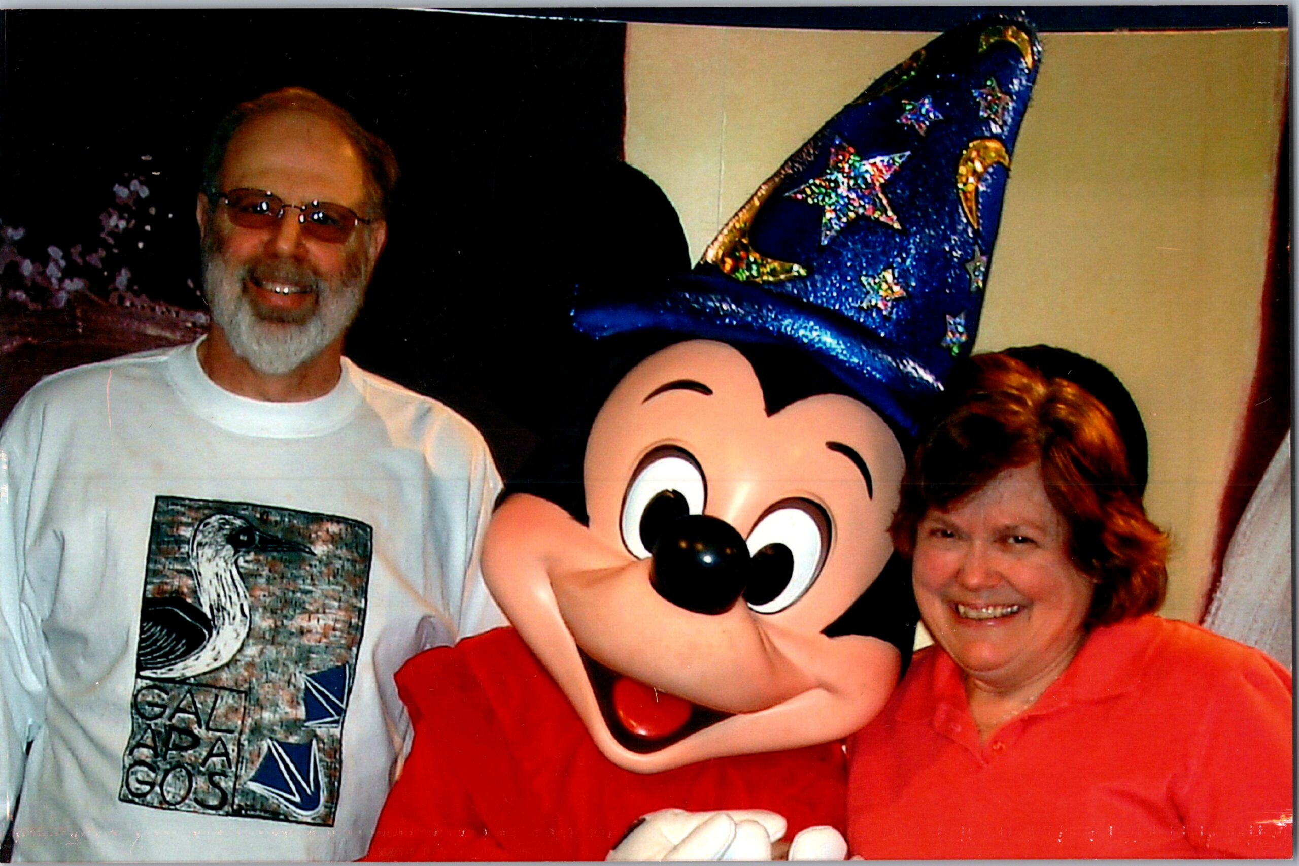 Three people pose with a life-size Mickey Mouse mascot: a bearded man in glasses on the left and a woman in red on the right.