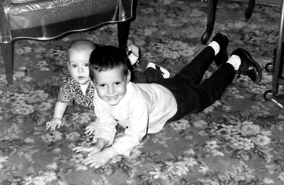Two young children crawl on a floral carpet, the older boy smiling at the camera while the baby watches nearby.