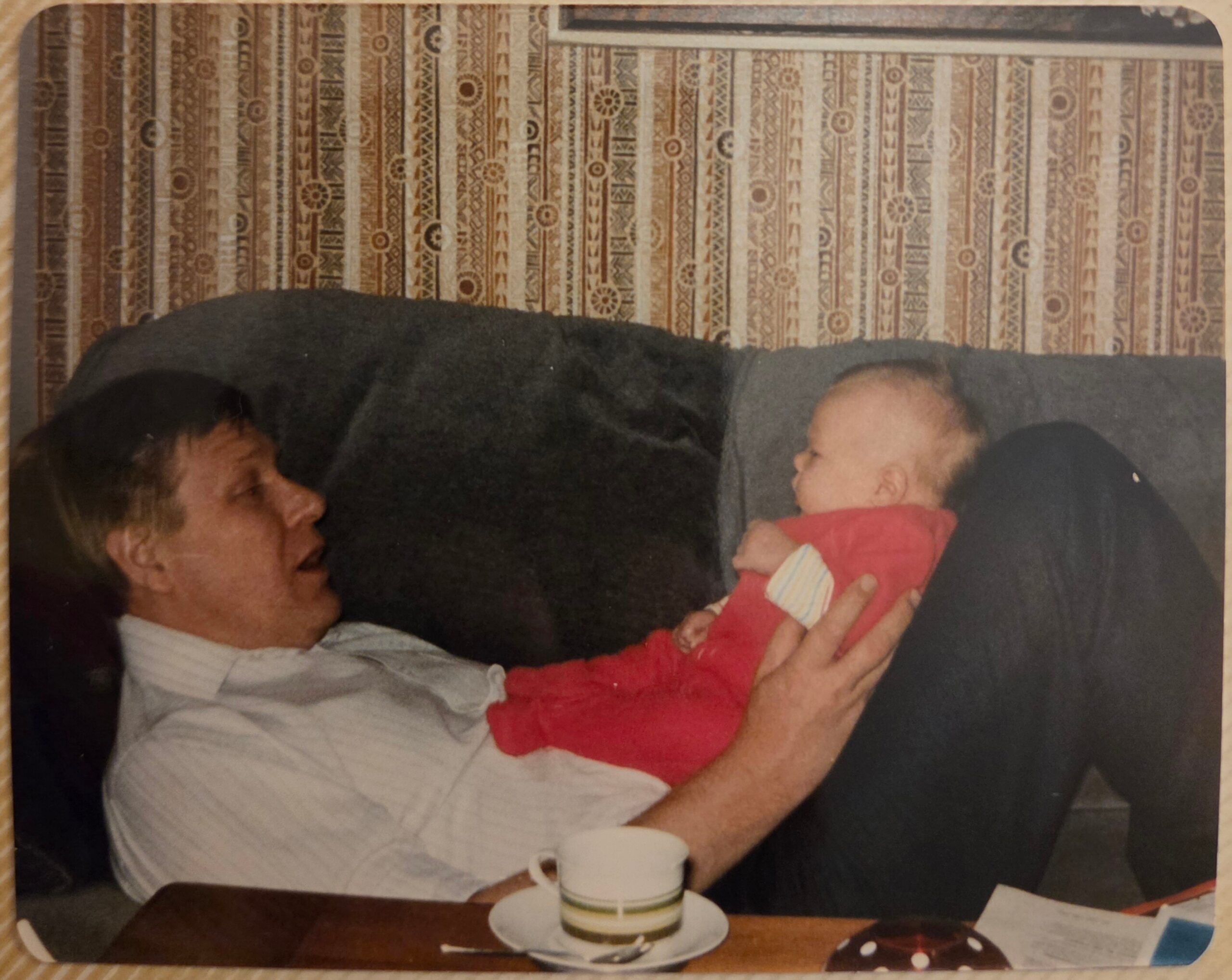 Man with gray hair sitting on a couch, holding a baby in a red outfit facing him, near a patterned wallpaper background and a coffee cup on the table in front.