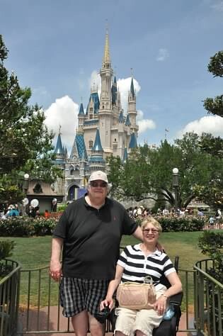 Older man and woman posing in front of Cinderella Castle at Disneyland, with greenery and a crowd in the background.