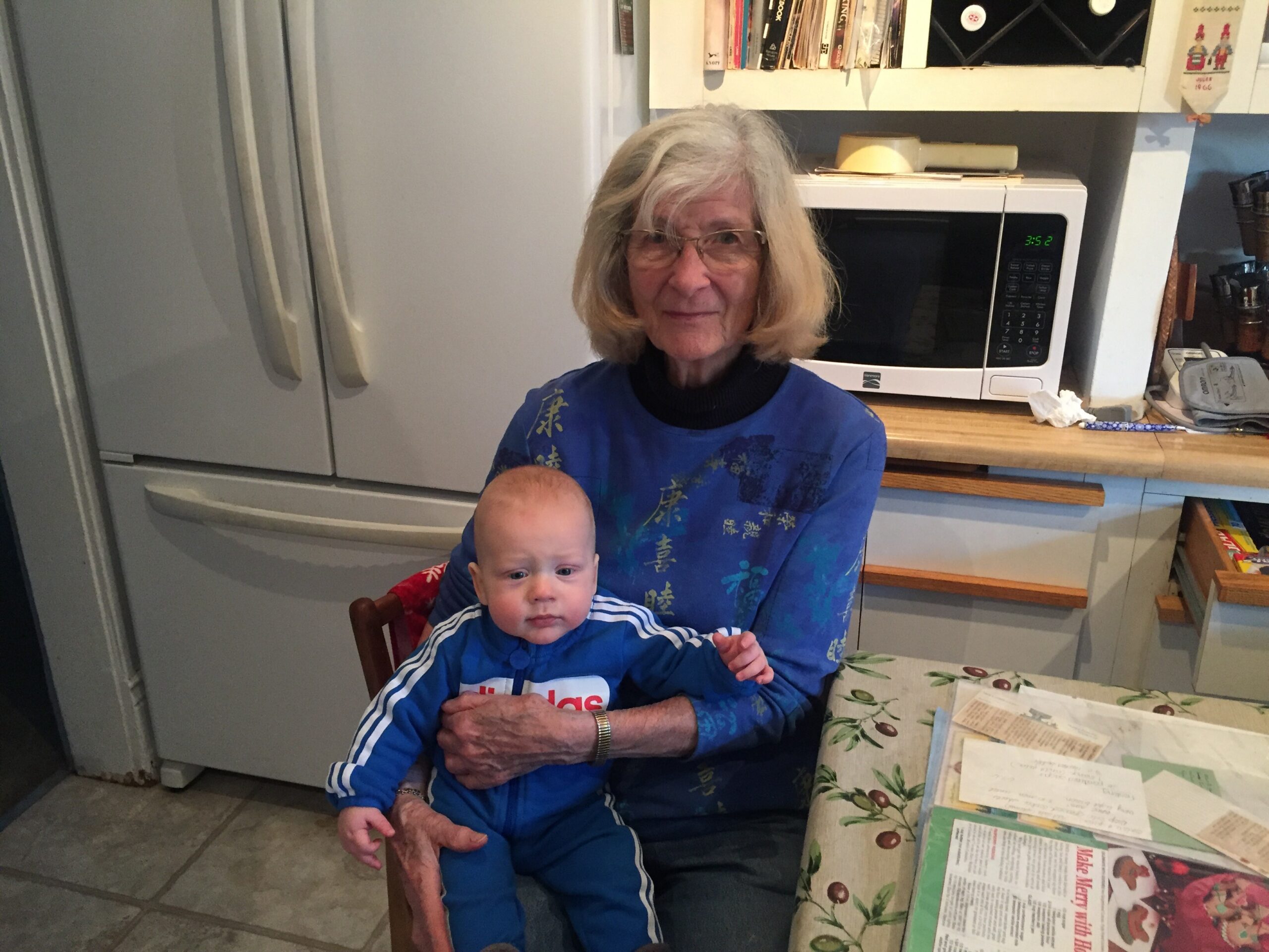 Older woman with gray hair wearing blue shirt sits in a kitchen, holding a baby on her lap while facing the camera.