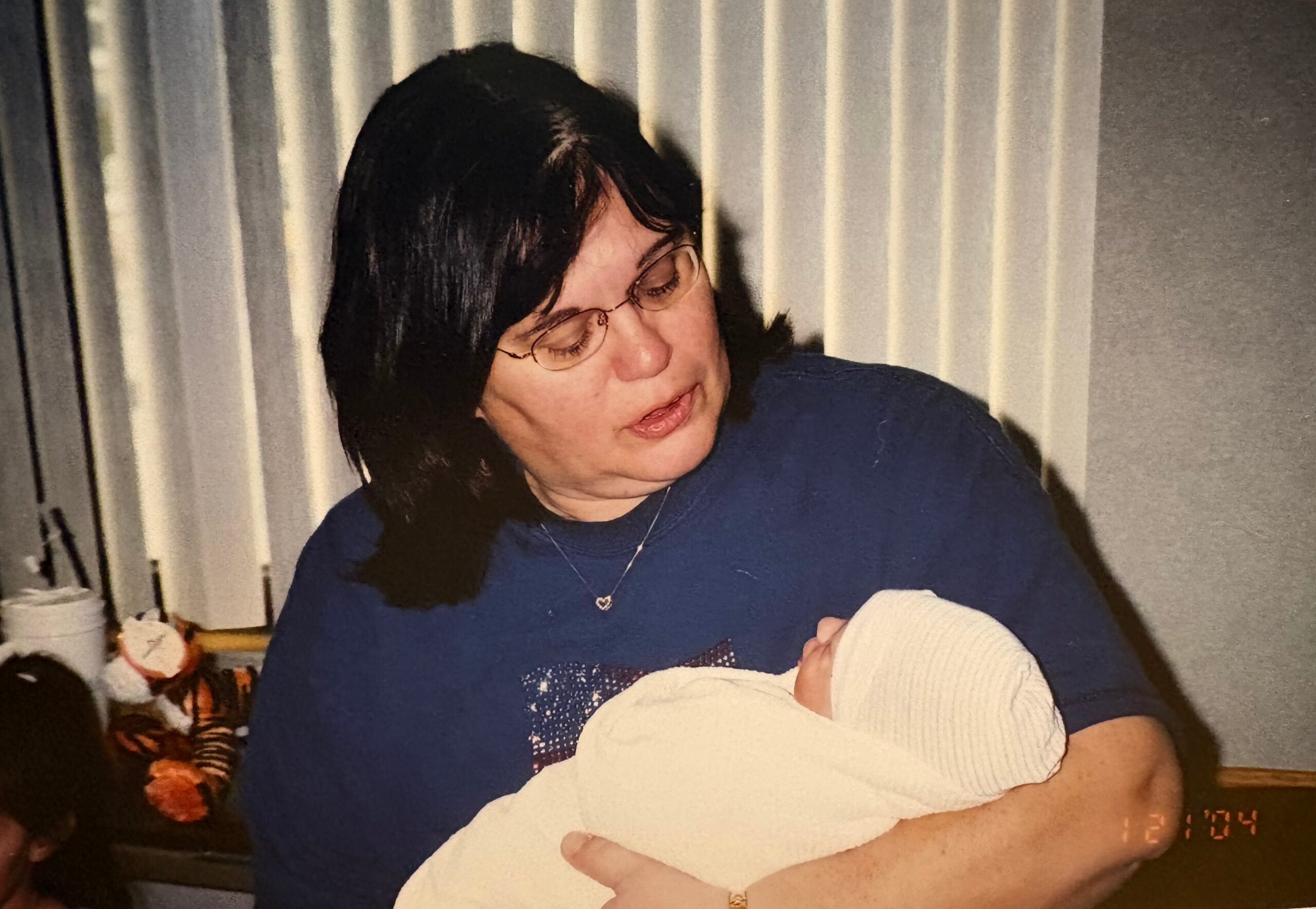 Woman with short dark hair and glasses cradling a newborn swaddled in a white blanket indoors.
