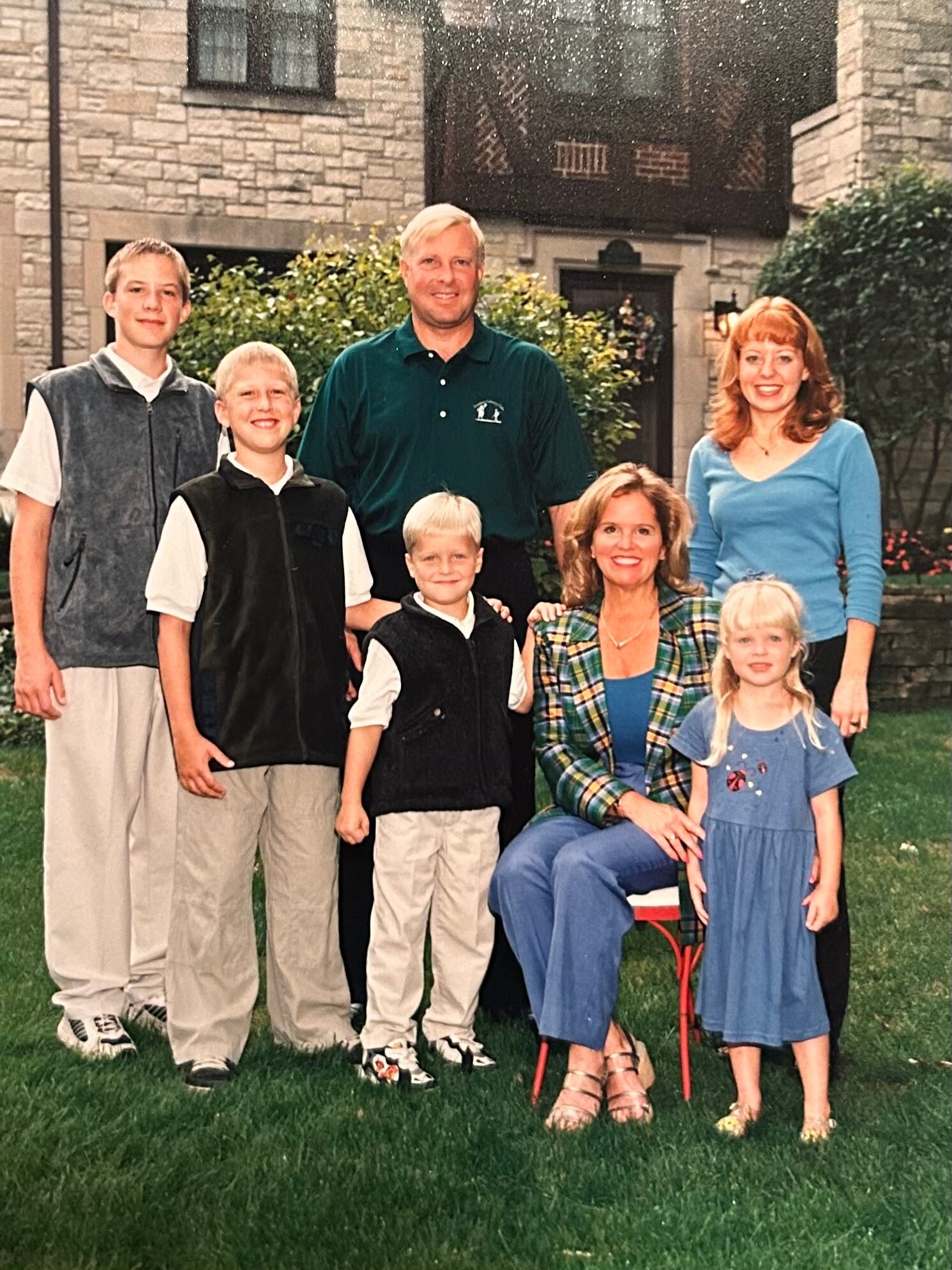 Family of seven posing for a photo in a front yard with a stone house in the background. A man in a green polo stands in the center; others stand or kneel around him, with a woman in plaid and a woman in blue on the right and several children in front.