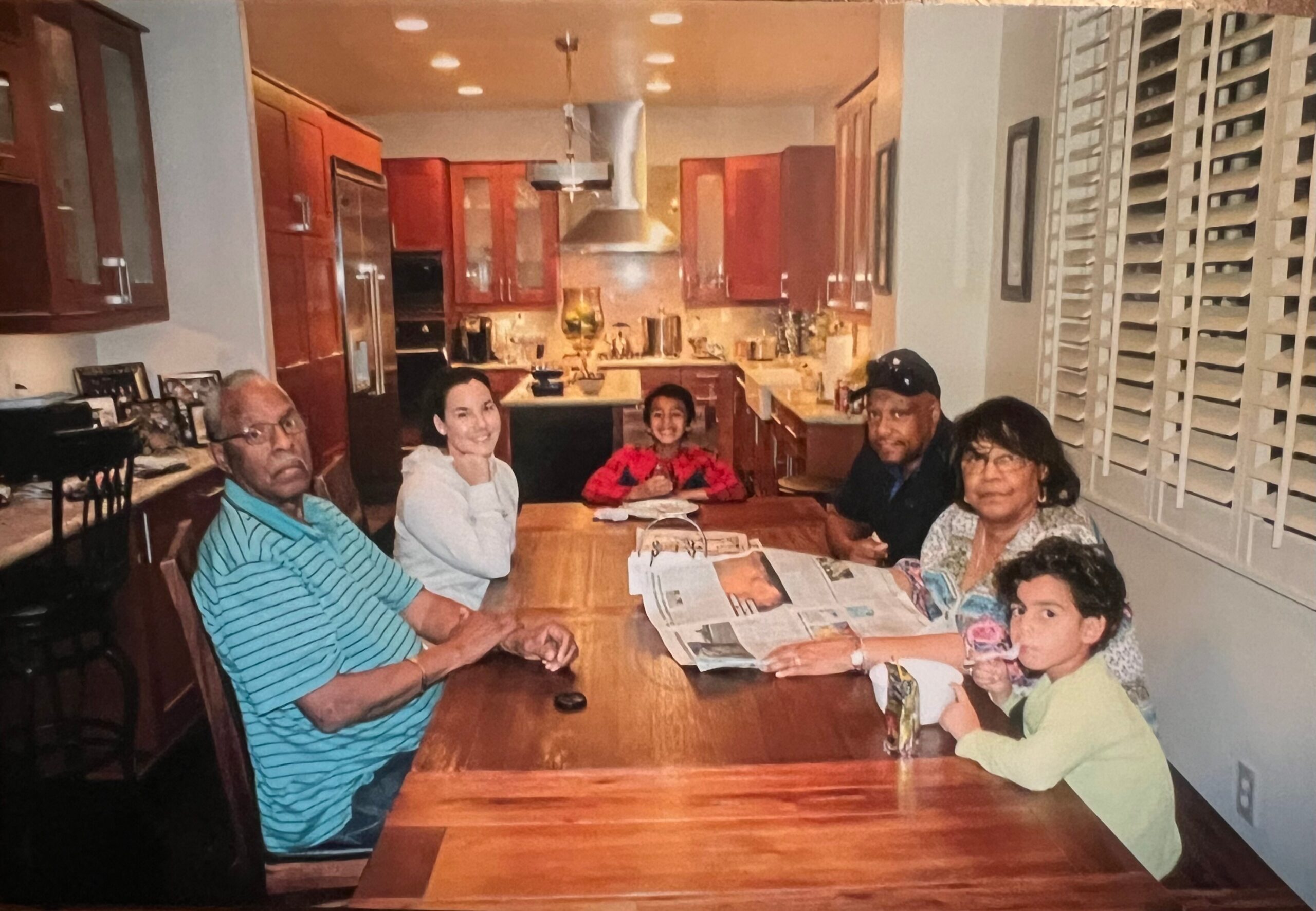 Multigenerational family sits around a wooden dining table in a kitchen, smiling at the camera.
