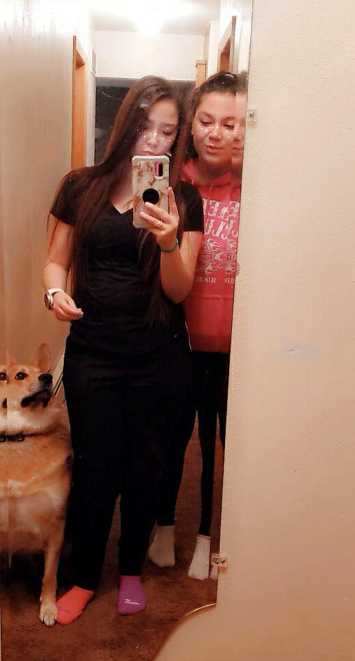 Two girls take a mirror selfie in a hallway, one in black and the other in a pink hoodie, with a dog sitting by their feet on the left.