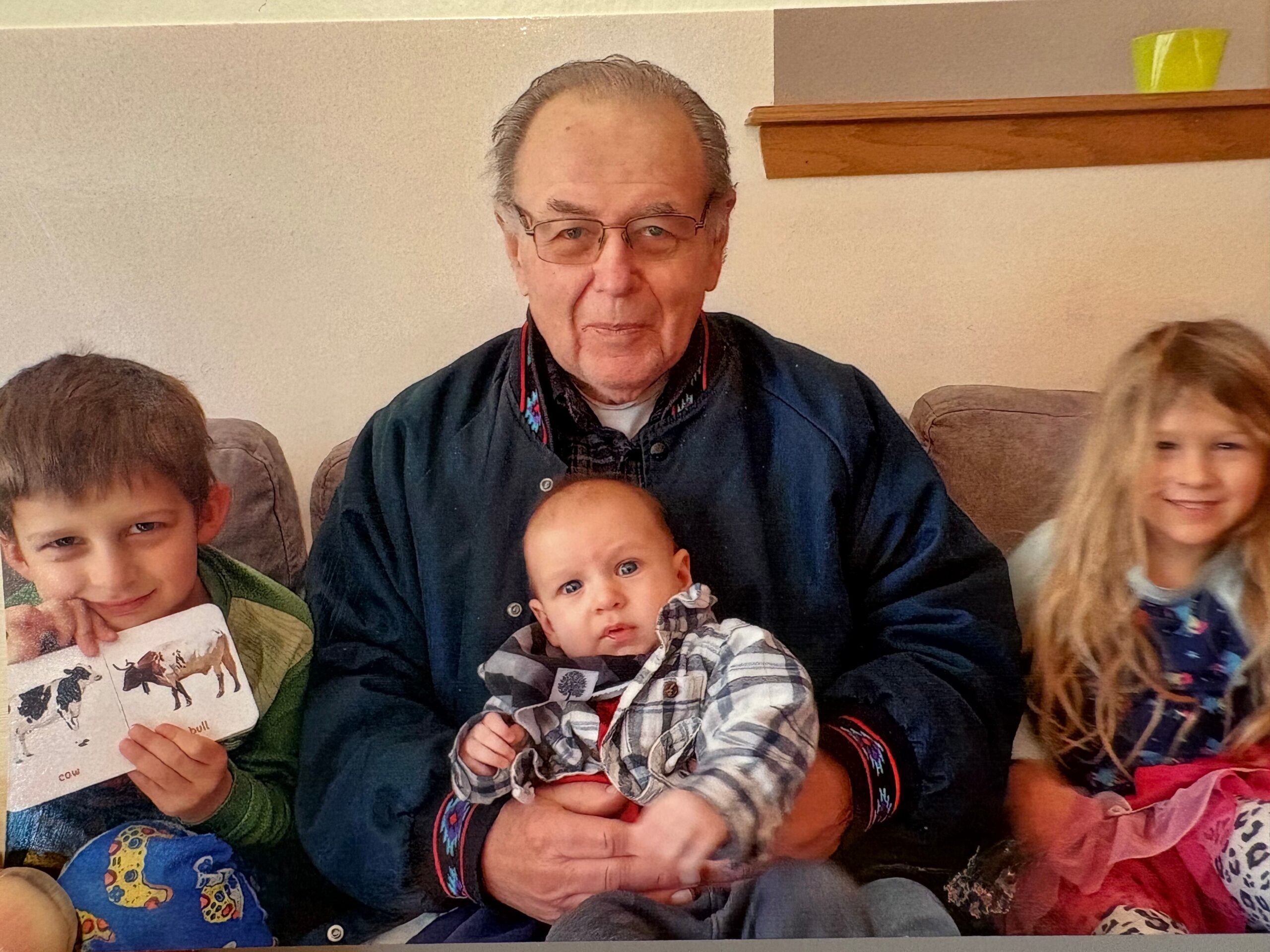 Older man wearing glasses sits on a couch, holding a baby while children flank him on either side.