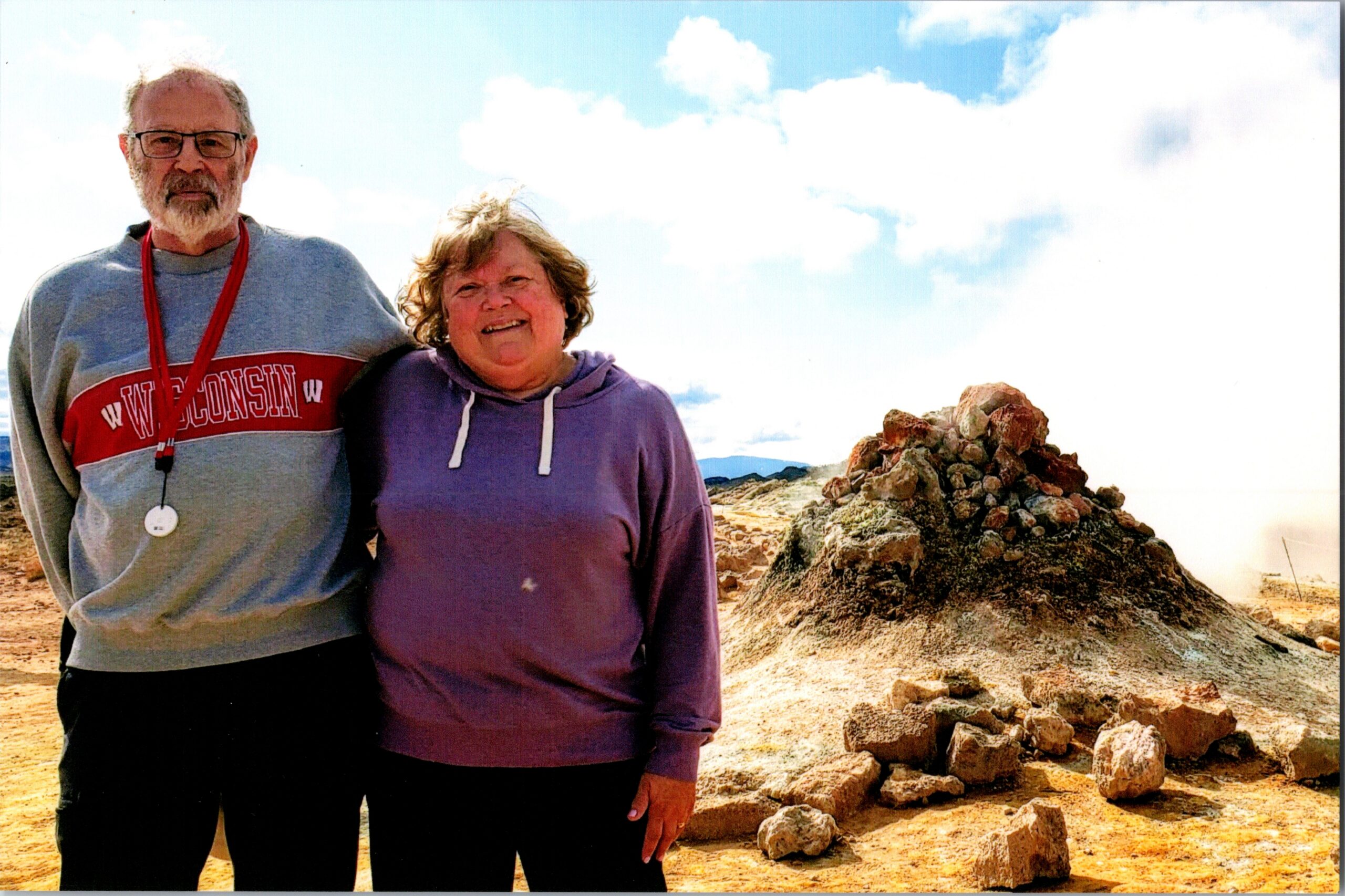 Two adults stand side by side outdoors on a rocky desert landscape under a cloudy blue sky.